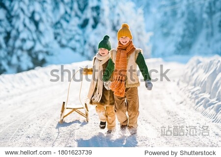 Two little kids have fun in the beautiful winter nature with snow-covered trees. Children walk along a snowy road pulling a sled. Winter knitted wool retro clothes.