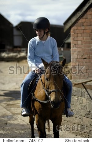 一个年轻的金发女孩骑着棕色马的附近农场建筑