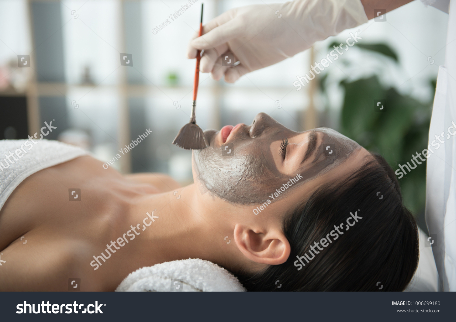 Side view of asian girl getting skin treatment at beauty salon. Beautician is holding brush near her face. Healthy facial mask concept