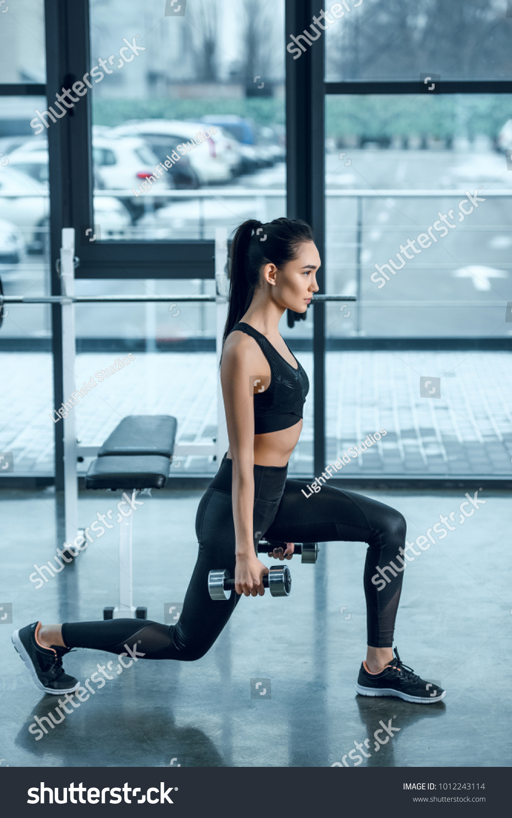side view of young fit woman doing single leg squats with dumbbells at gym