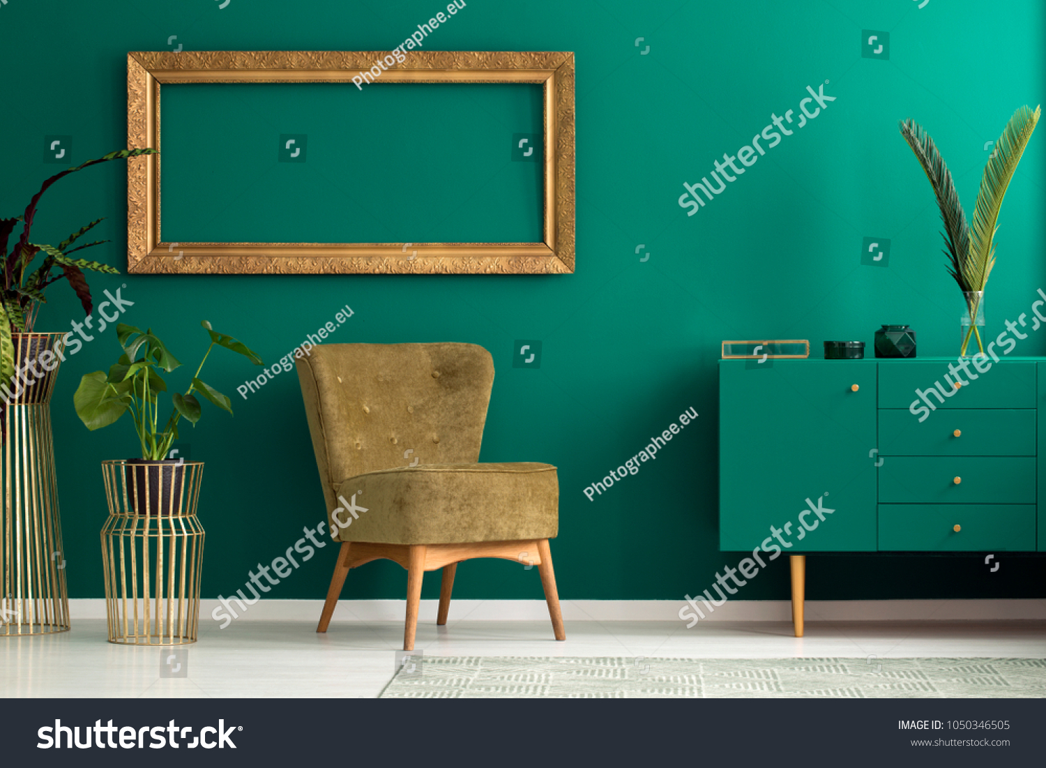 Palm leaf on a modern teal sideboard with drawers in a luxurious green living room interior with golden decorations and an upholstered chair