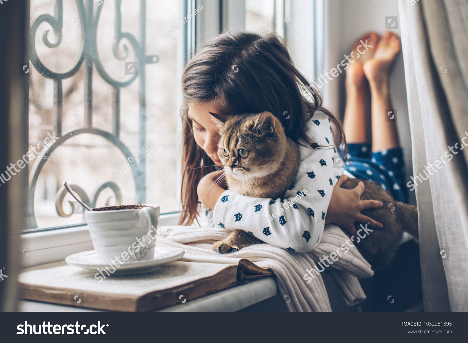 Child in pajamas relaxing on a window sill with pet. Lazy weekend with cat at home. Cozy scene hygge concept.