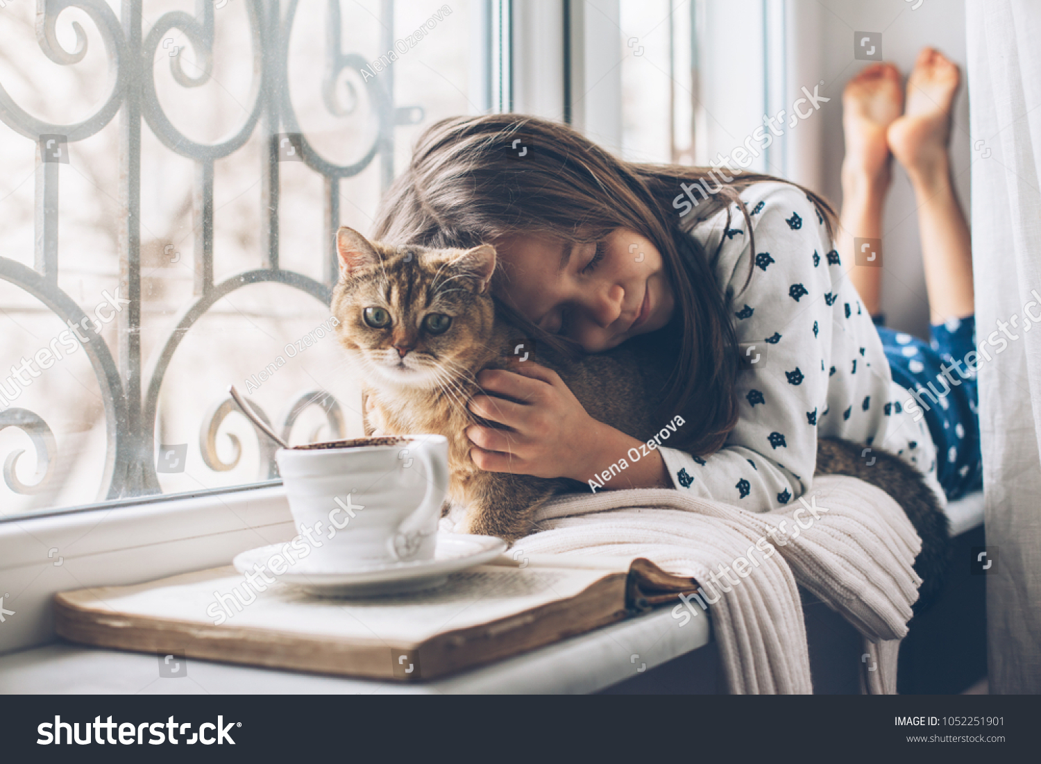 Child in pajamas relaxing on a window sill with pet. Lazy weekend with cat at home. Cozy scene hygge concept.