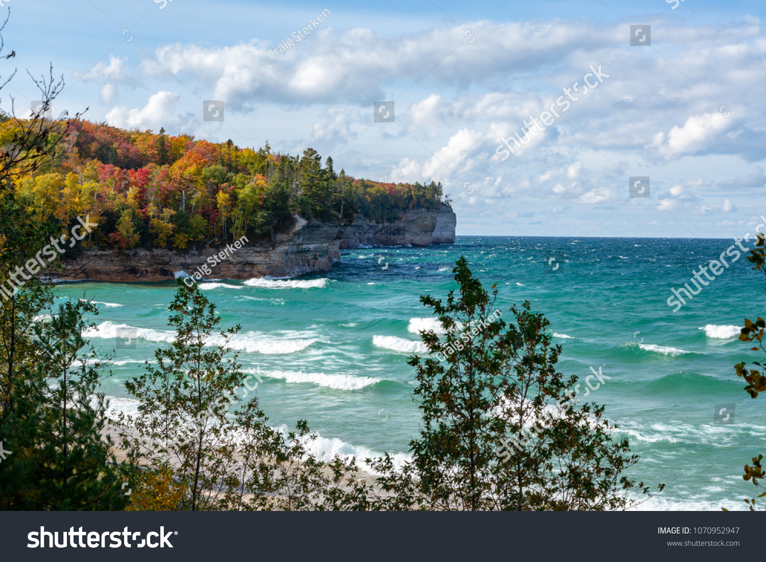 An Upper Peninsula forest creates an autumn background at Chapel Beach in northern Michigan. Lake Superior waves crash on the beach and white puffy clouds dot the blue sky.