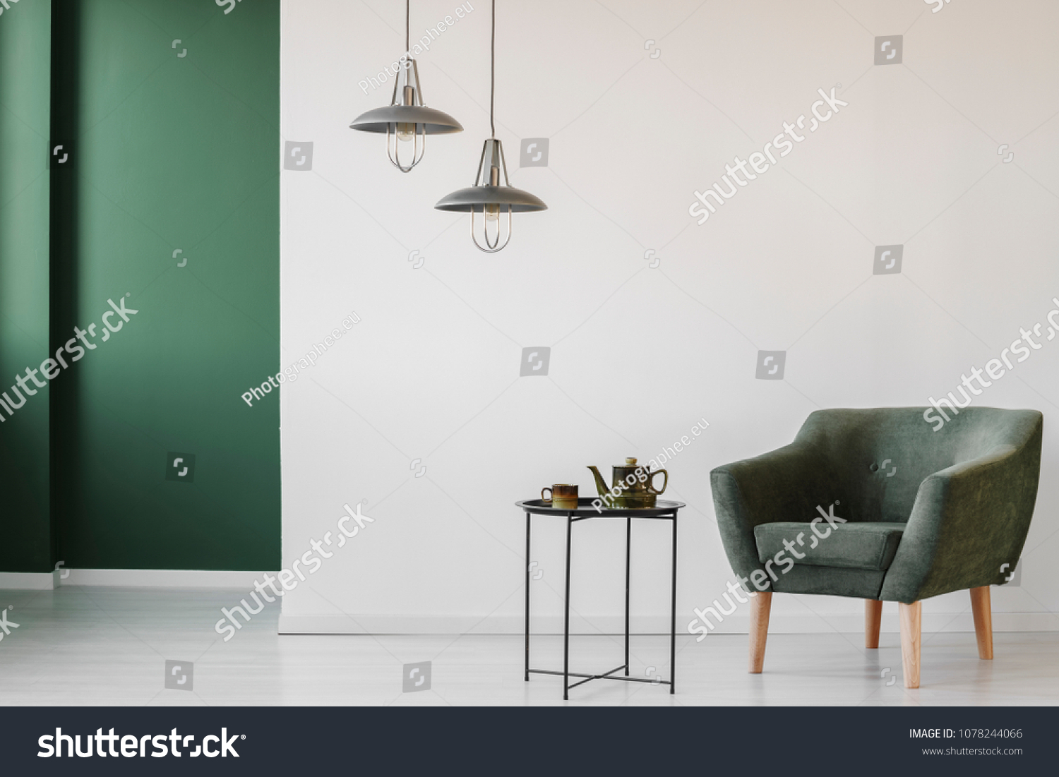 Upholstered dark armchair and an industrial side table with a tea kettle and cup in a minimalist living room interior with a green corner