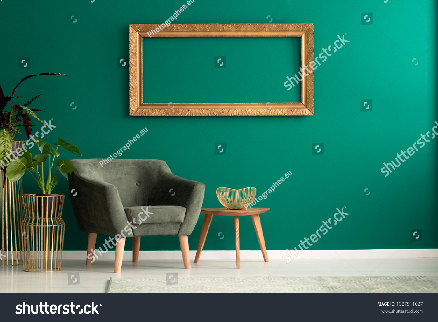 Wooden table with gold metal bowl standing by a green armchair in living room interior with frame on the wall