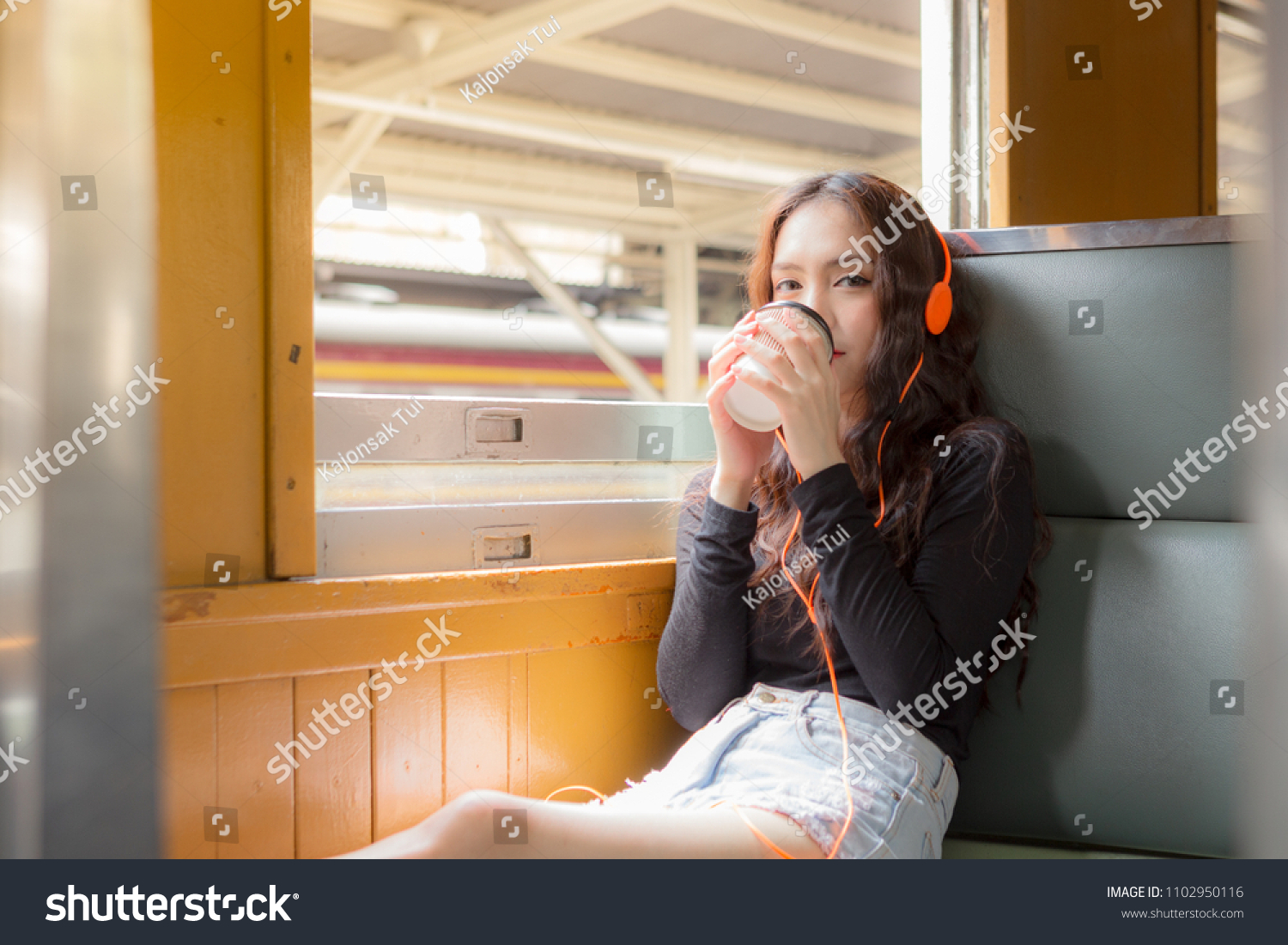 A beautiful charming asian girl drinking a delicious coffee while listening music with headphone. Gorgeous woman sit near window in retro train in morning. Feeling happy & relax. Portrait lady travel.