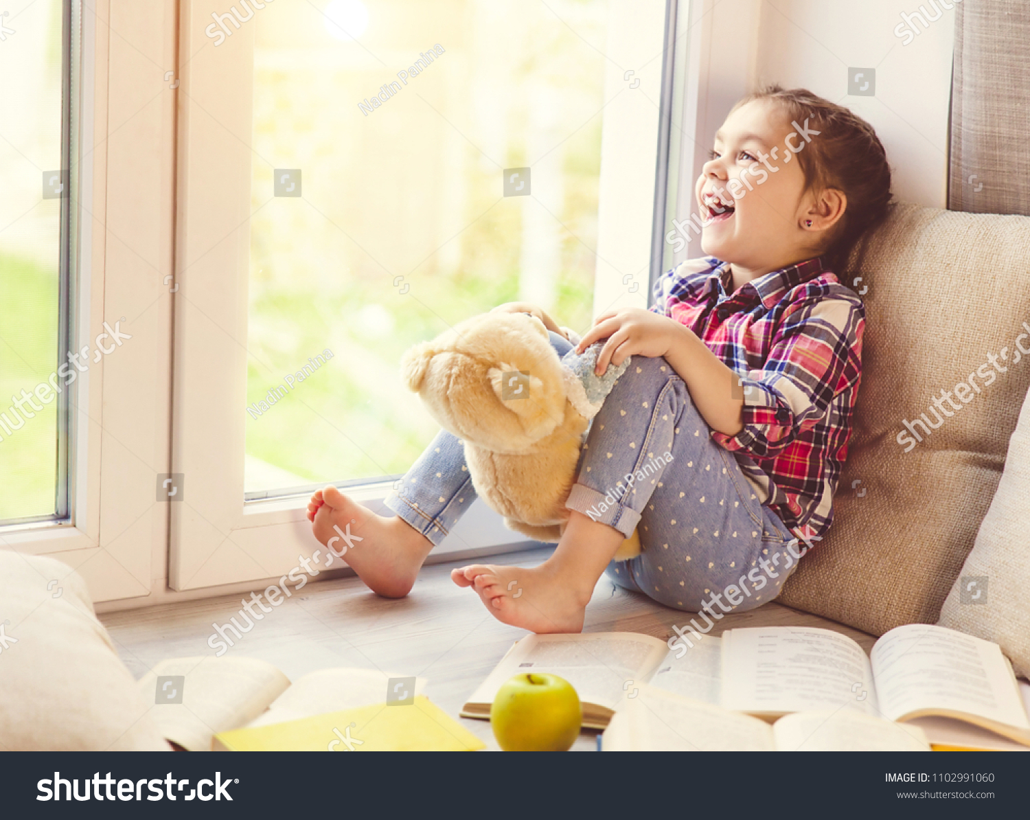 Cute little toddler girl sitting by the window with her teddy bear and laughing.