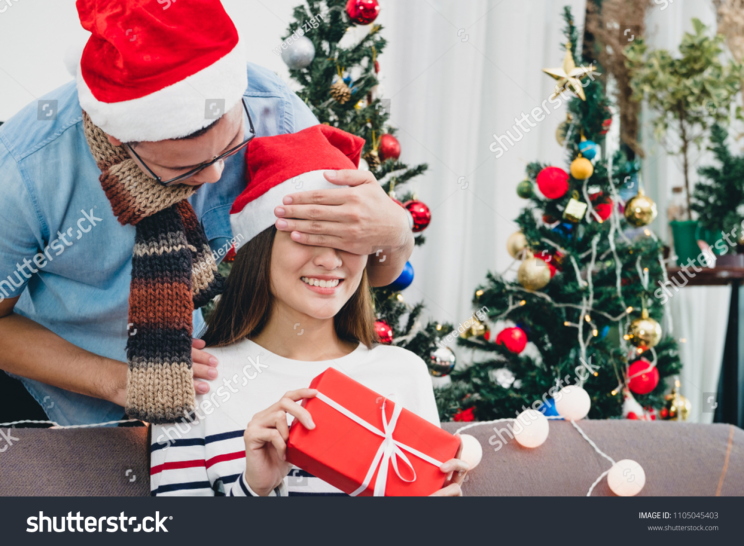 Asia lovers couple boyfriend surprise girlfriendby giving Christmas present at sofa with xmas decoration tree at house party Holiday festive celebrating season