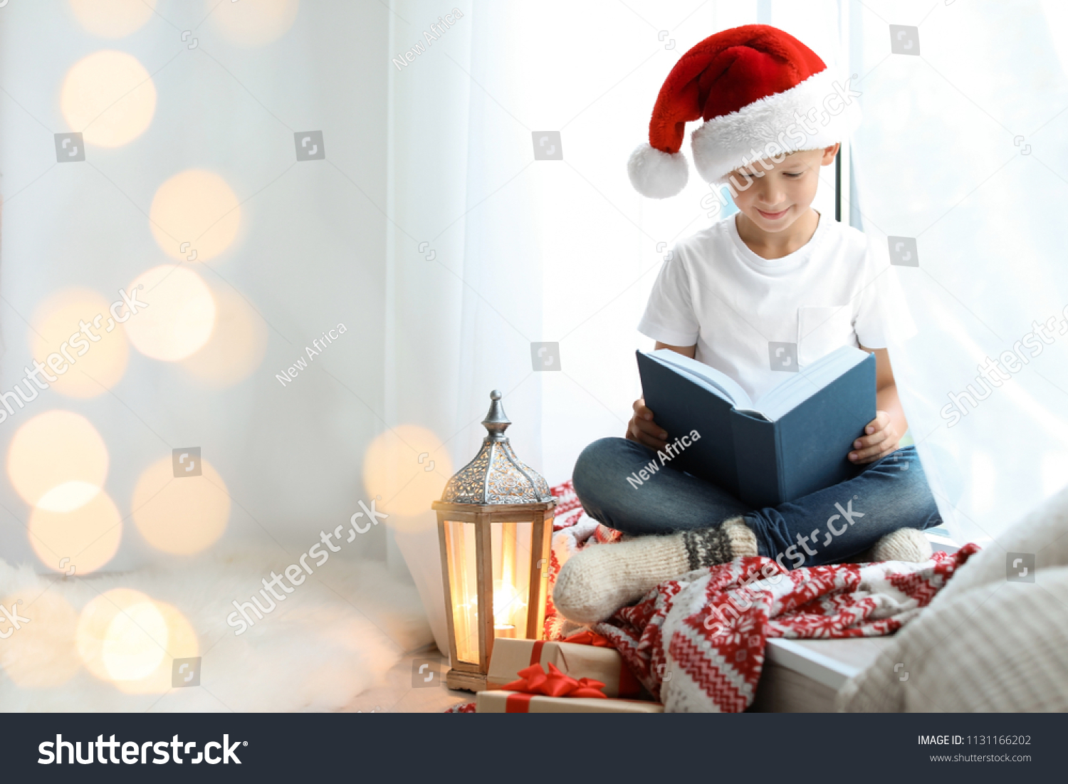 Cute little child in Santa hat reading Christmas story while sitting on windowsill at home