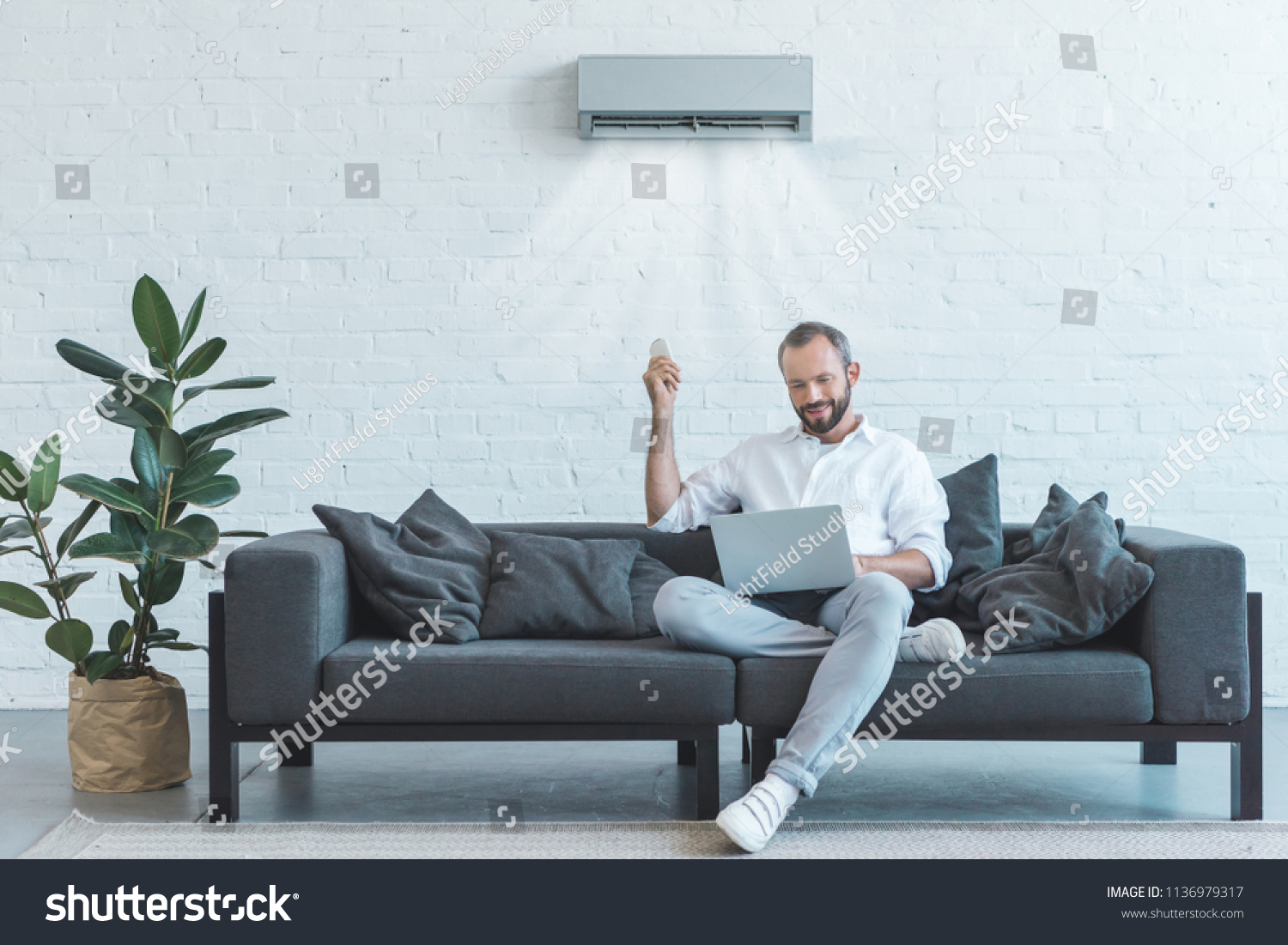 handsome man turning on air conditioner with remote control while using laptop on sofa at home