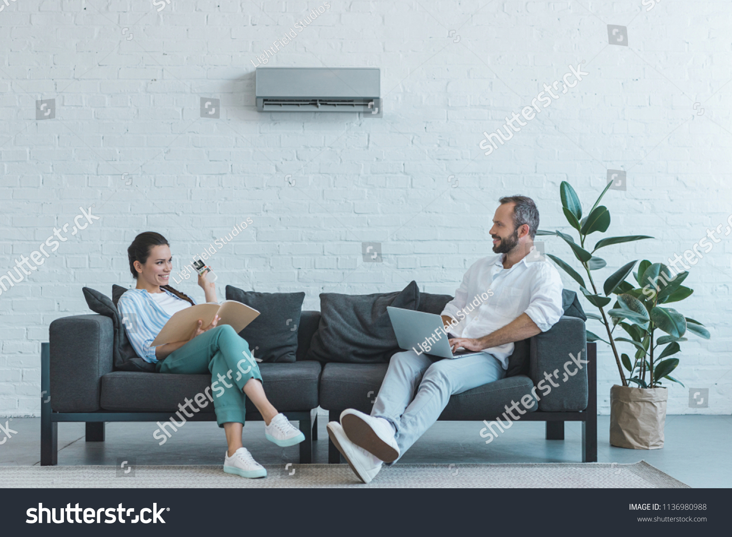 couple turning on air conditioner during the summer heat while sitting on sofa with book and laptop