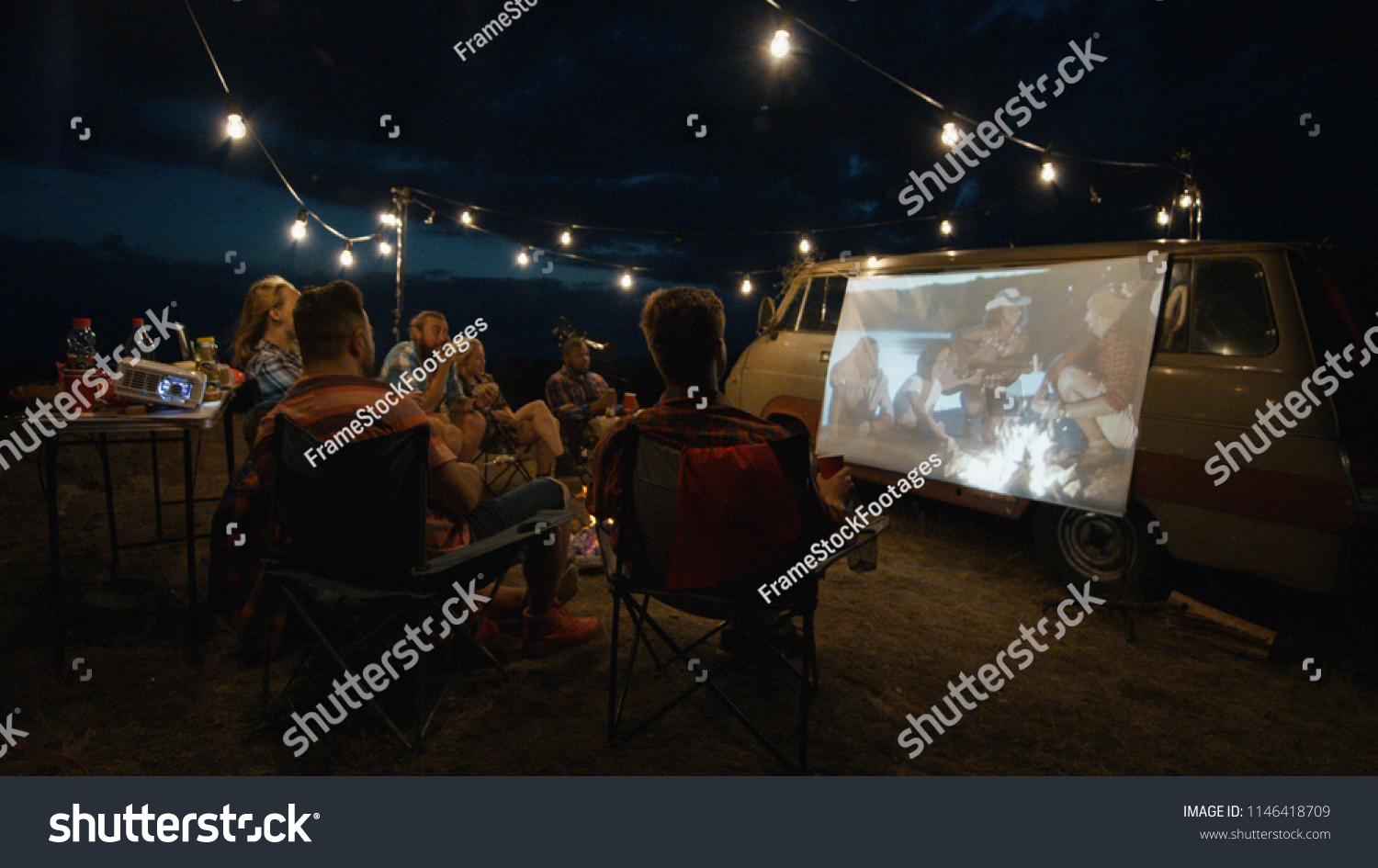 Friends gathering in campsite around bonfire and watching movie with projector on van side in dark evening