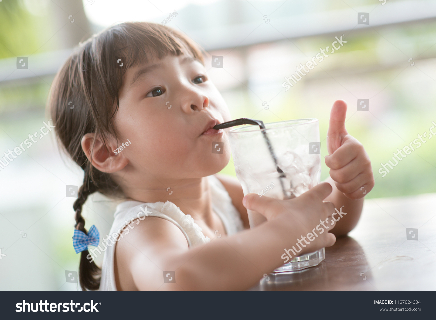 Little Asian child drinking ice chocolate and giving thumb up at cafe. Natural light outdoor lifestyle.