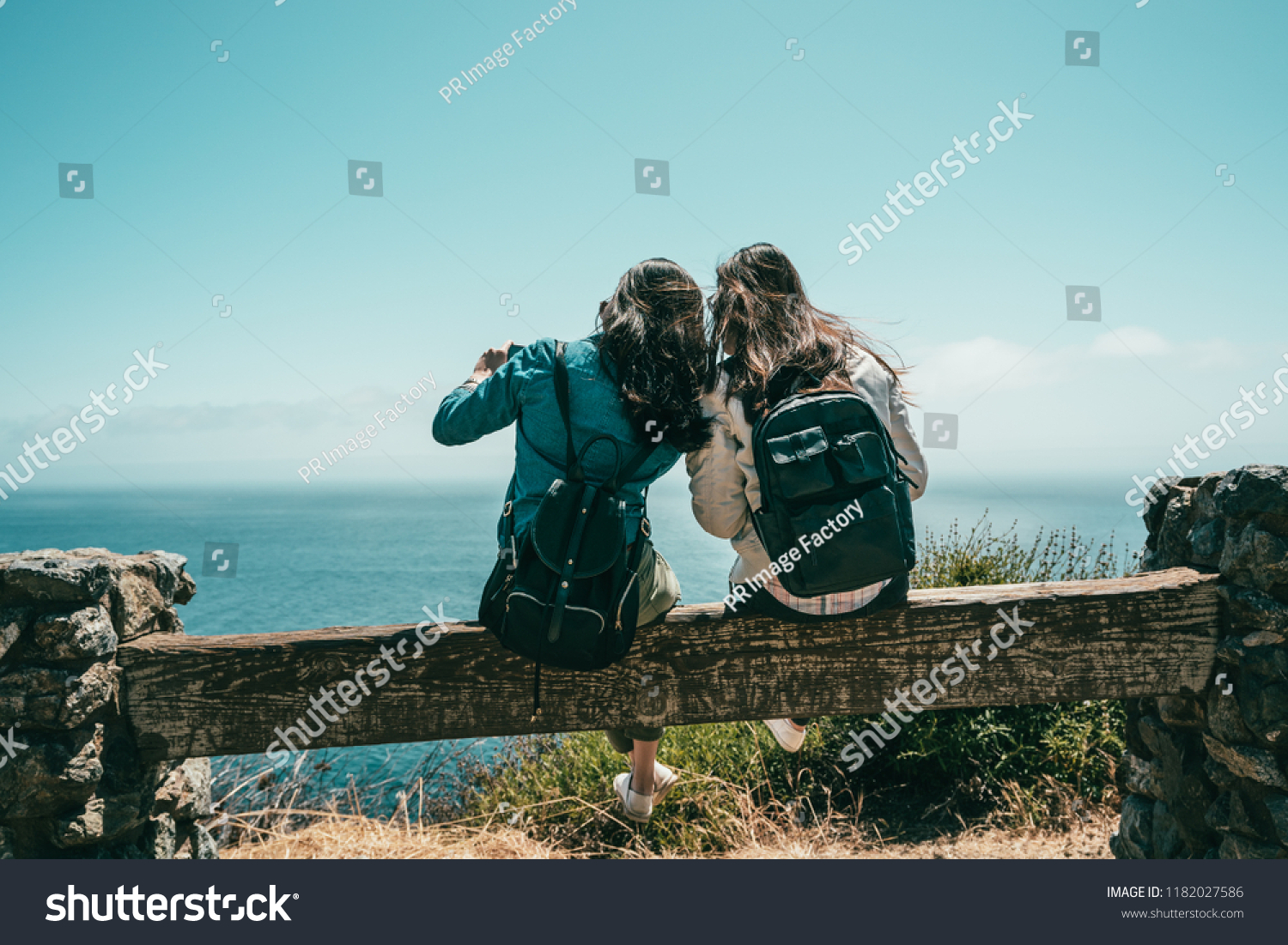 the back view of young good friends sitting together on the bench with a background of the stunning sea.