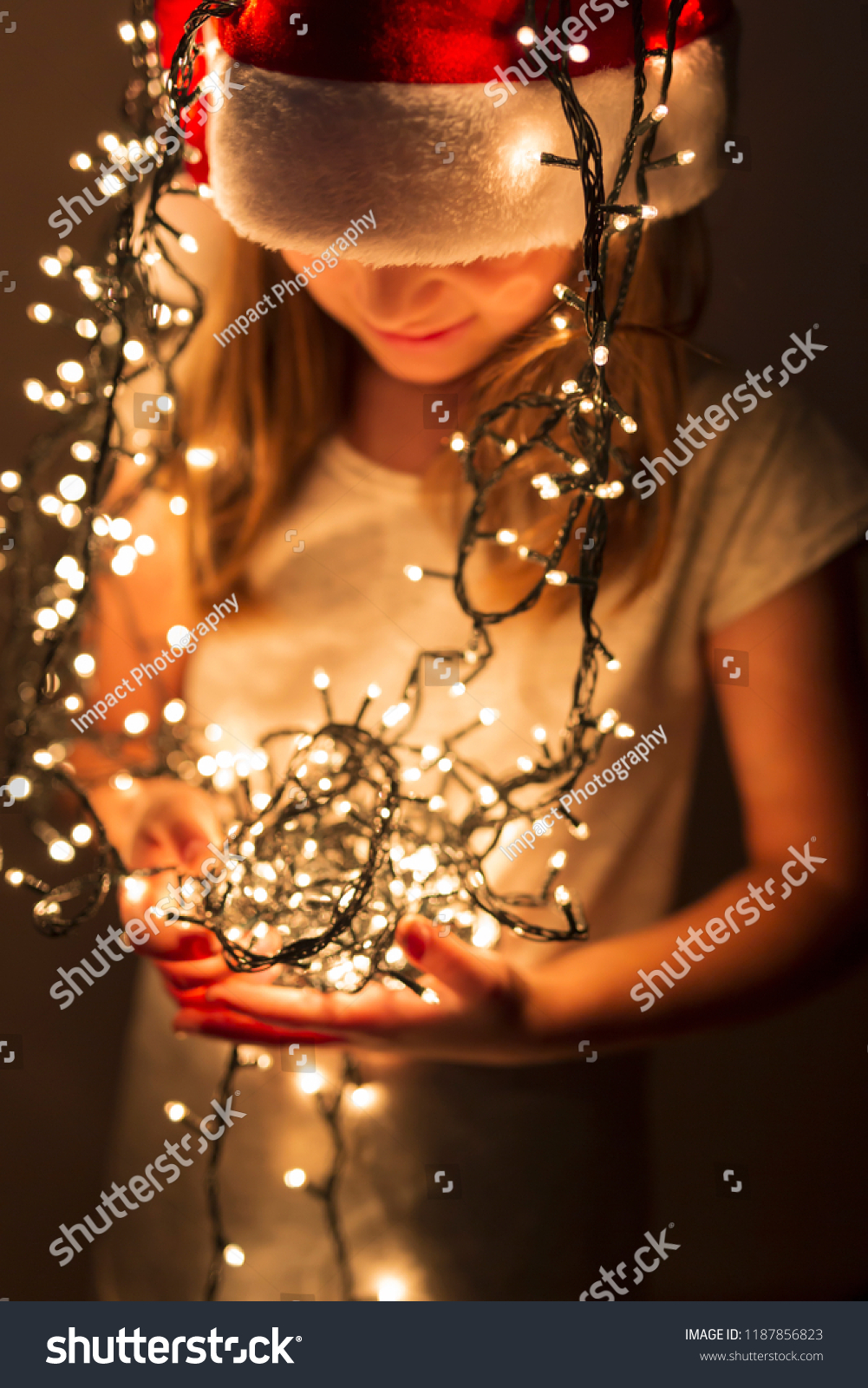Beautiful little girl wearing Santa hat and holding bunch of Christmas lights; child setting up Christmas decorations and waiting for Santa