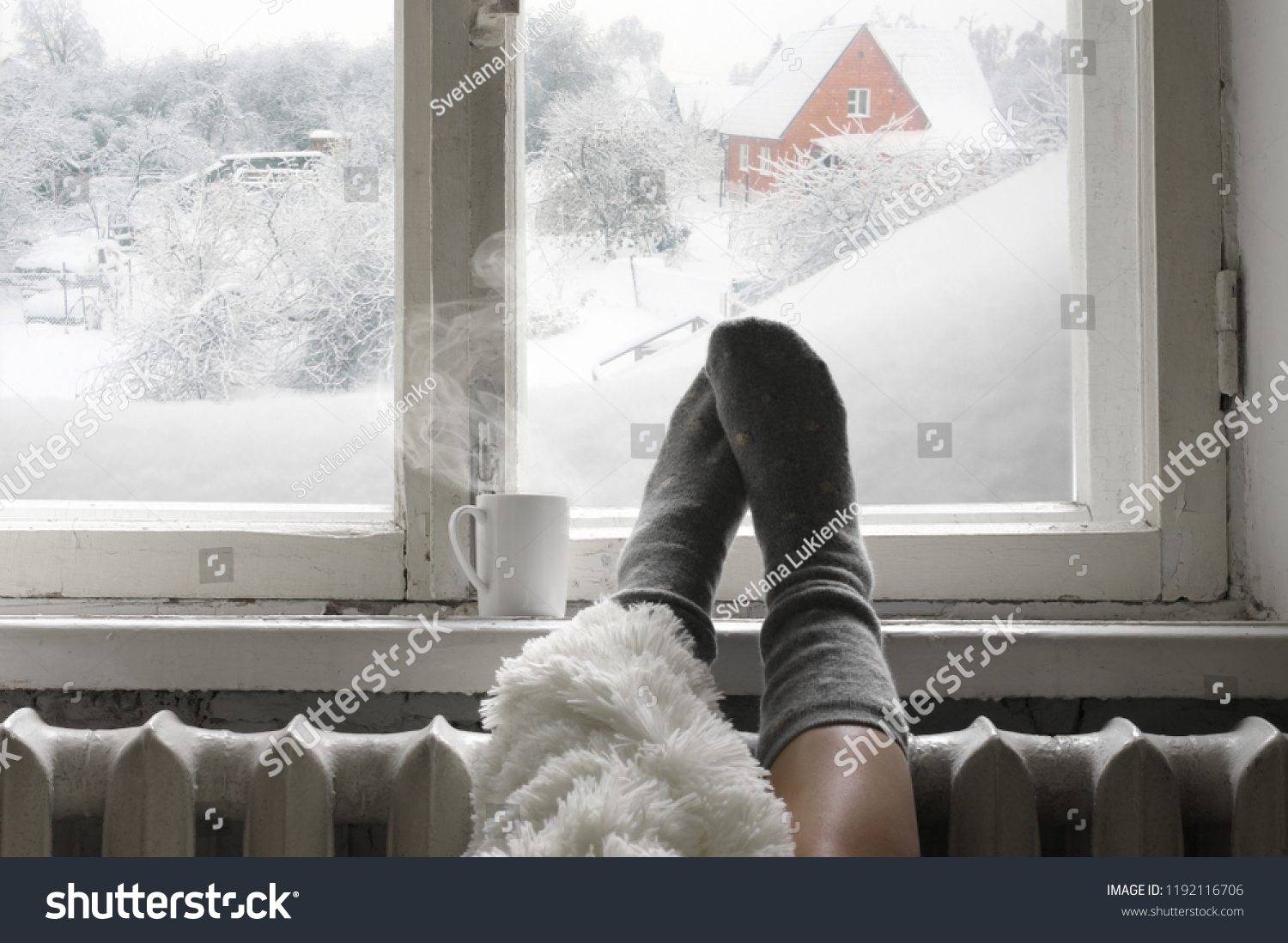 Cozy winter still life: woman legs in warm woolen socks under shaggy blanket and mug of hot beverage on old windowsill against snow landscape from outside.