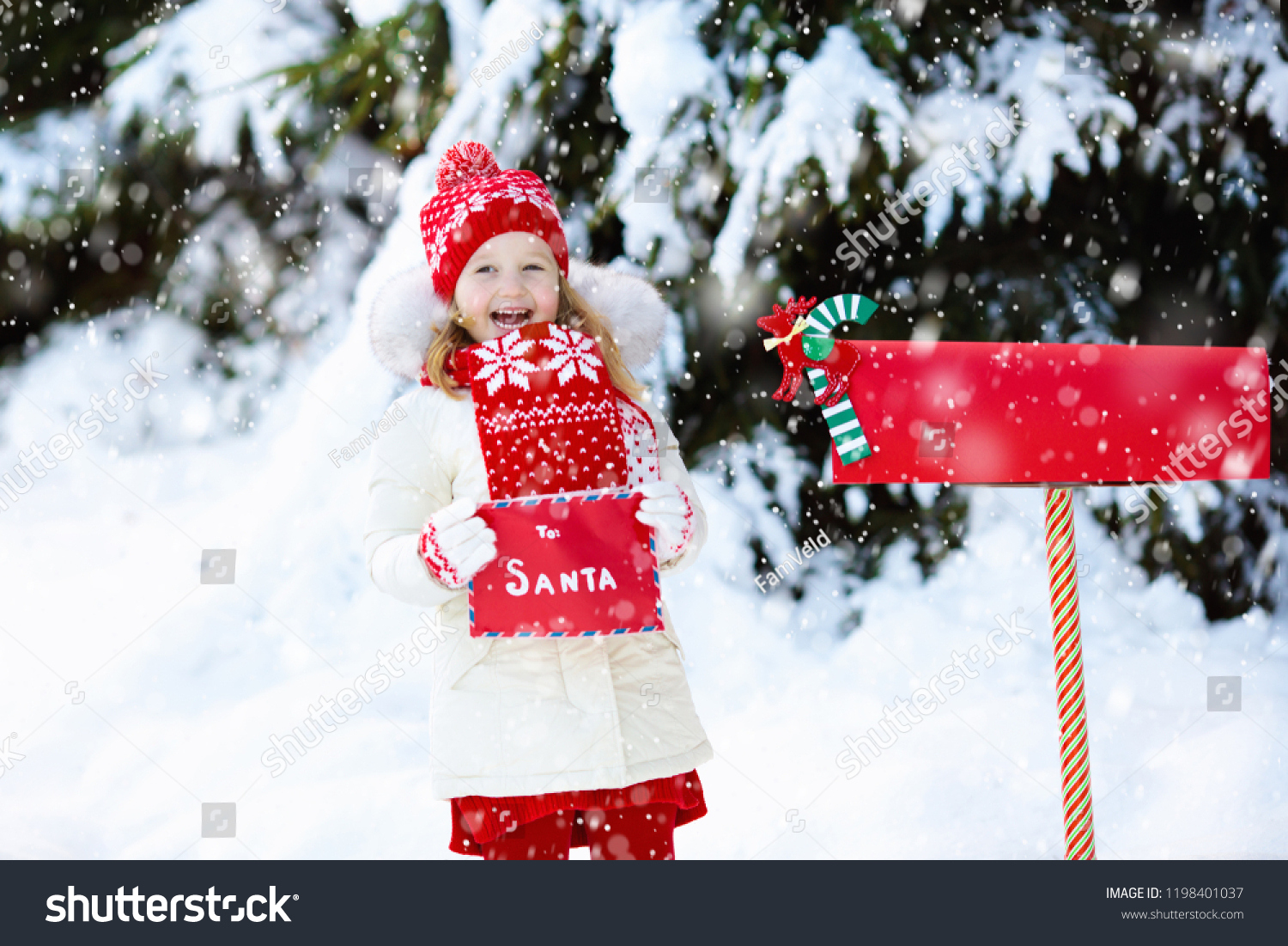 Happy child in knitted reindeer hat and scarf holding letter to Santa with Christmas presents wish list at red mail box in snow under Xmas tree in winter forest. Kids sending post to North Pole.