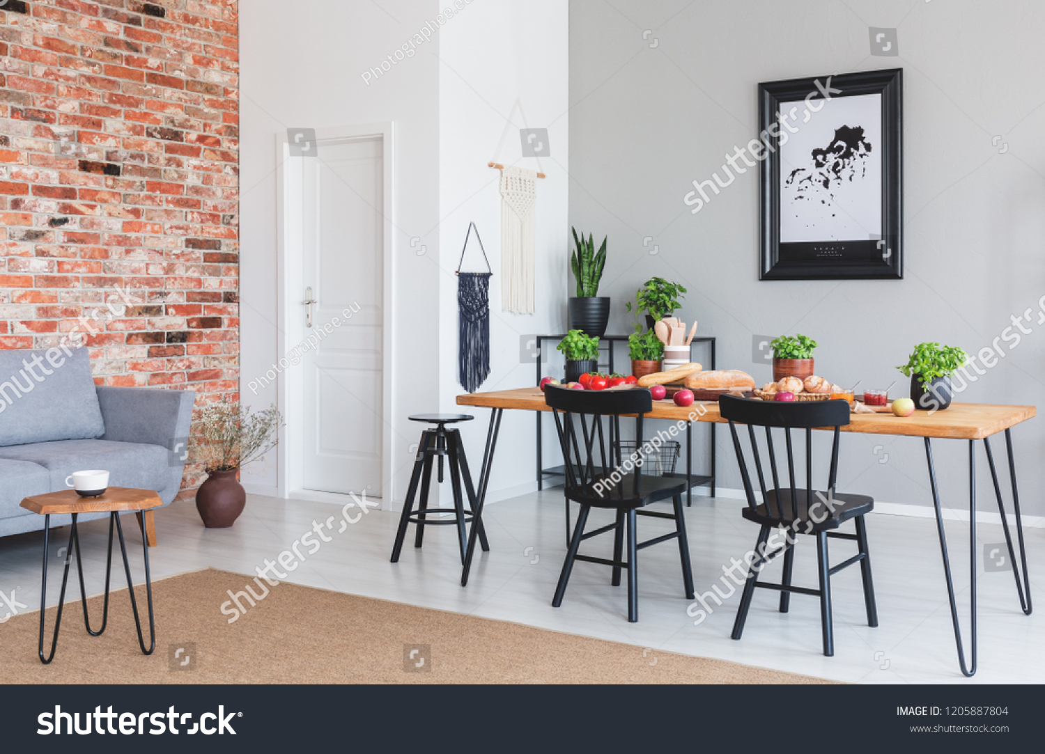 Black chairs at dining table and poster in flat interior with grey sofa against red brick wall. Real photo