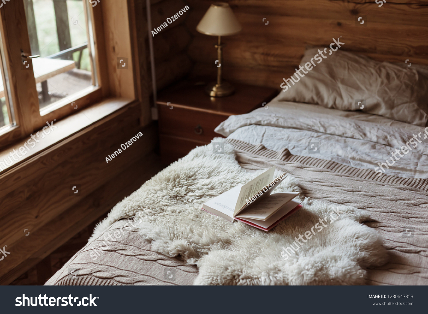Rustic interior of log cabin bedroom with bed by big window. Opened book on sheep rug. Warm and cozy weekend morning in hotel.