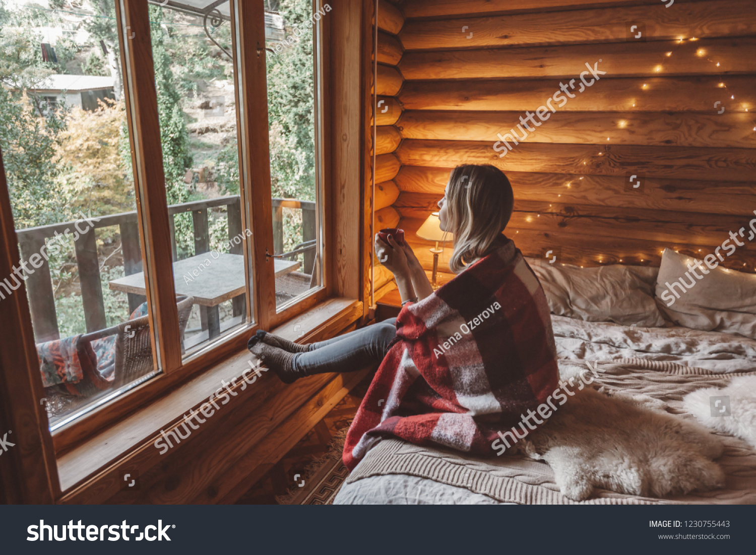 Woman in warm blanket relaxing and drinking morning coffee on cozy bed in log cabin in winter