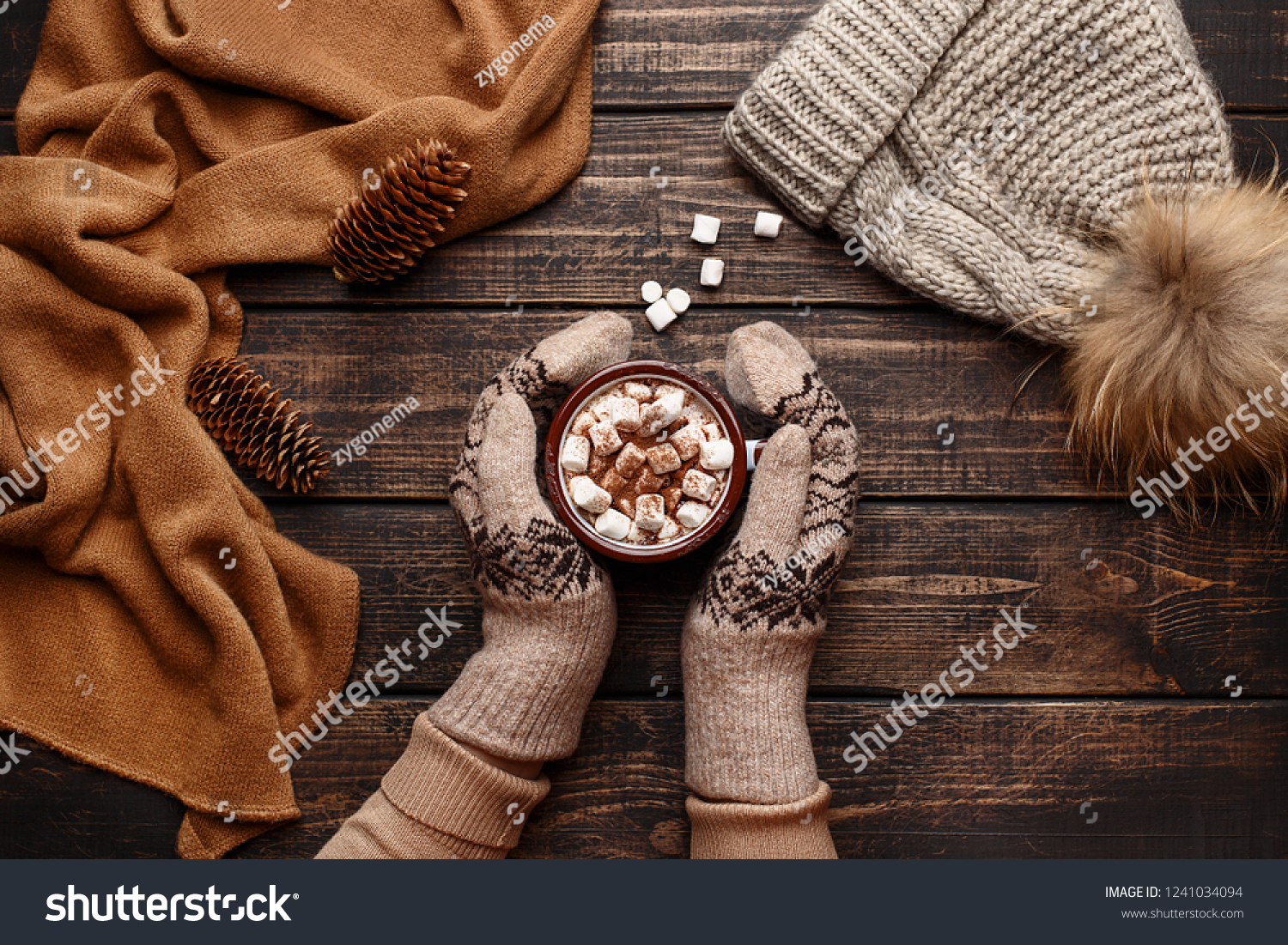 Winter flat lay with scarf knitted hat and female hands in mitten holding mug with hot chocolate or cocoa with marshmallow on dark rustic wooden table. Christmas and new year background. Top view