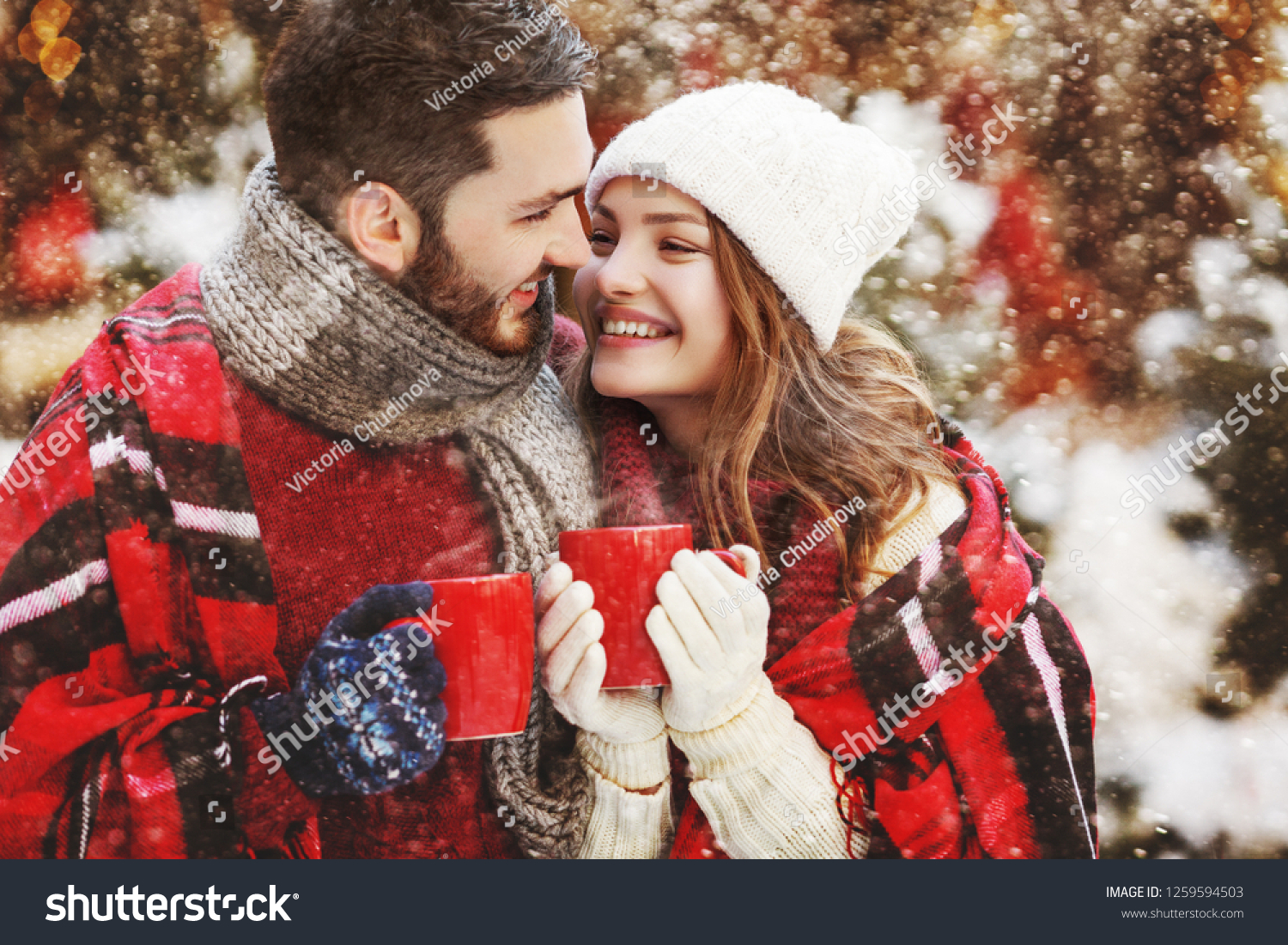 Young beautiful happy smiling couple holding red cups mugs of hot drink. Models looking at each other bundled up tartan blanket. Winter holidays Christmas celebration winter holidays concept
