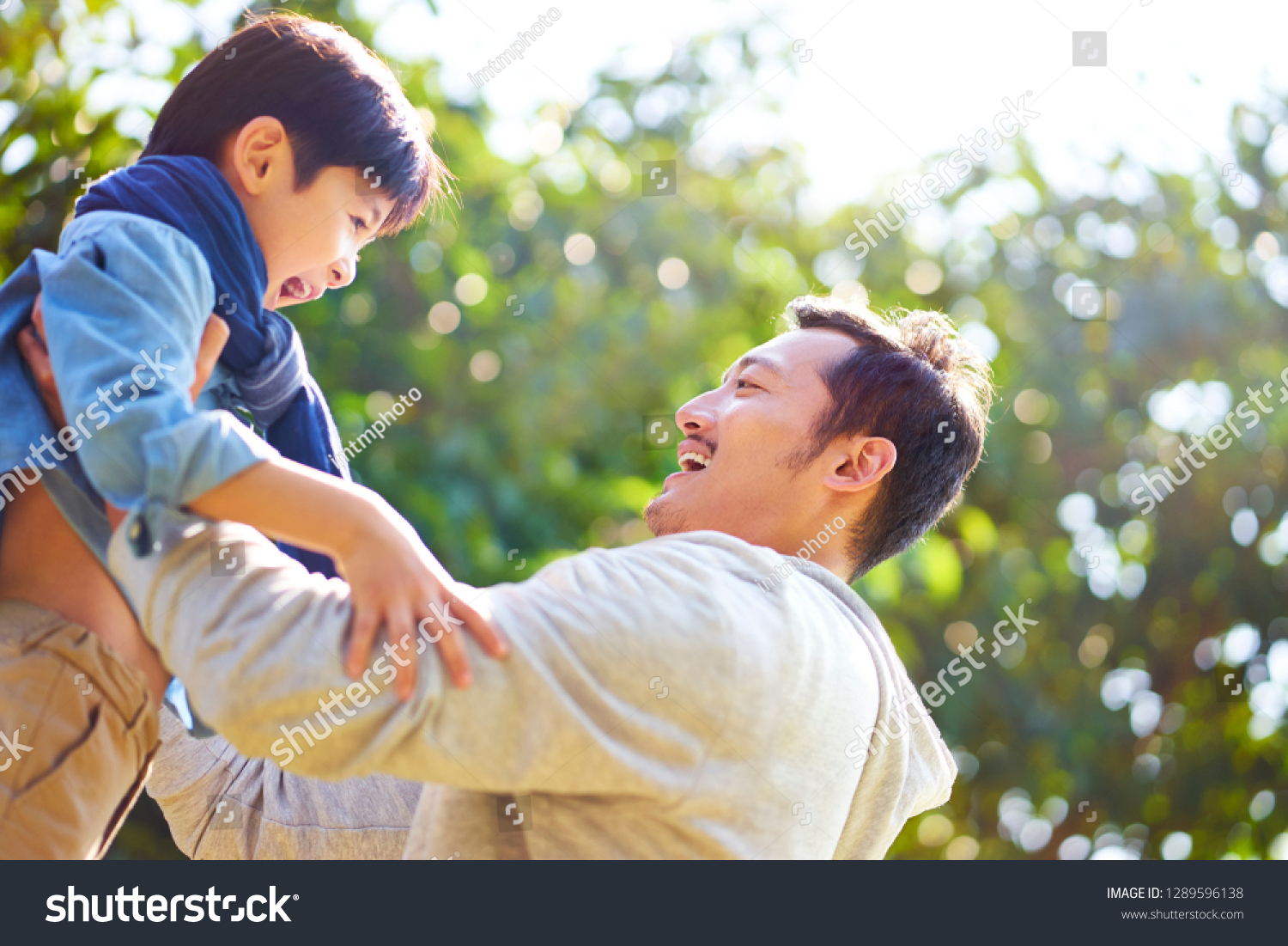 asian father having fun lifting son oudoors in park.