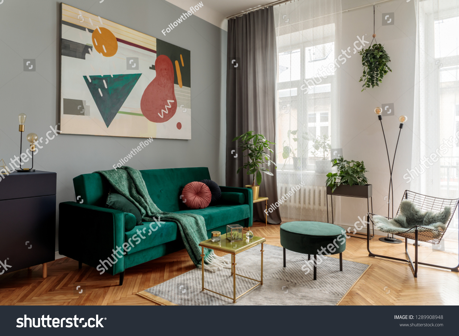Luxury and stylish interior with green velvet design sofa armchair tables and pouf. Tropical plants in design stand. Grey walls with abstract painting. Stylish decor of sitting room. Big windows.