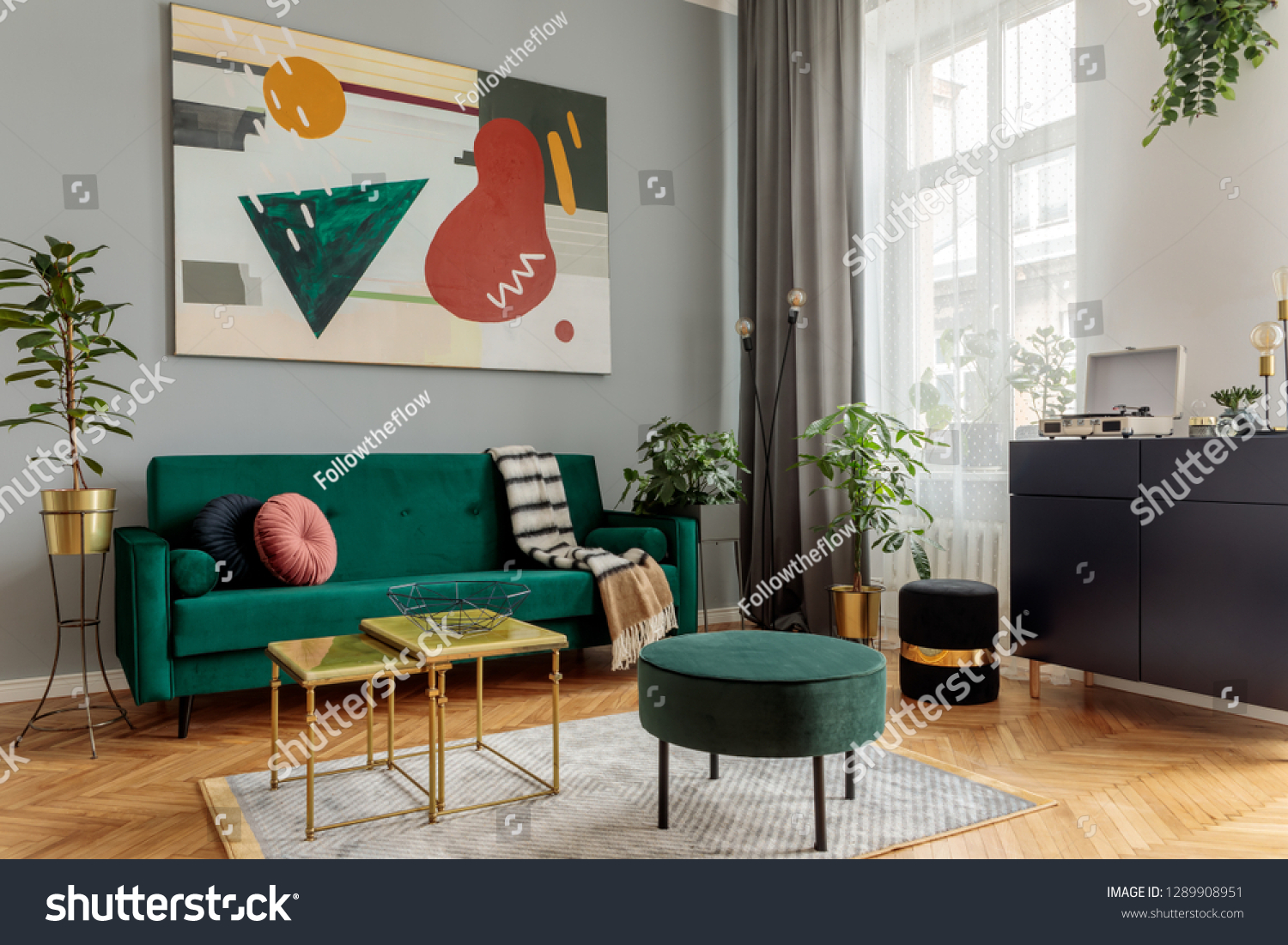 Luxury and modern home interior with design green sofa navy commode tables pouf and accessroies. A lot of plants in the room. Abstract painting. Stylish decor of living room with brown parquet.