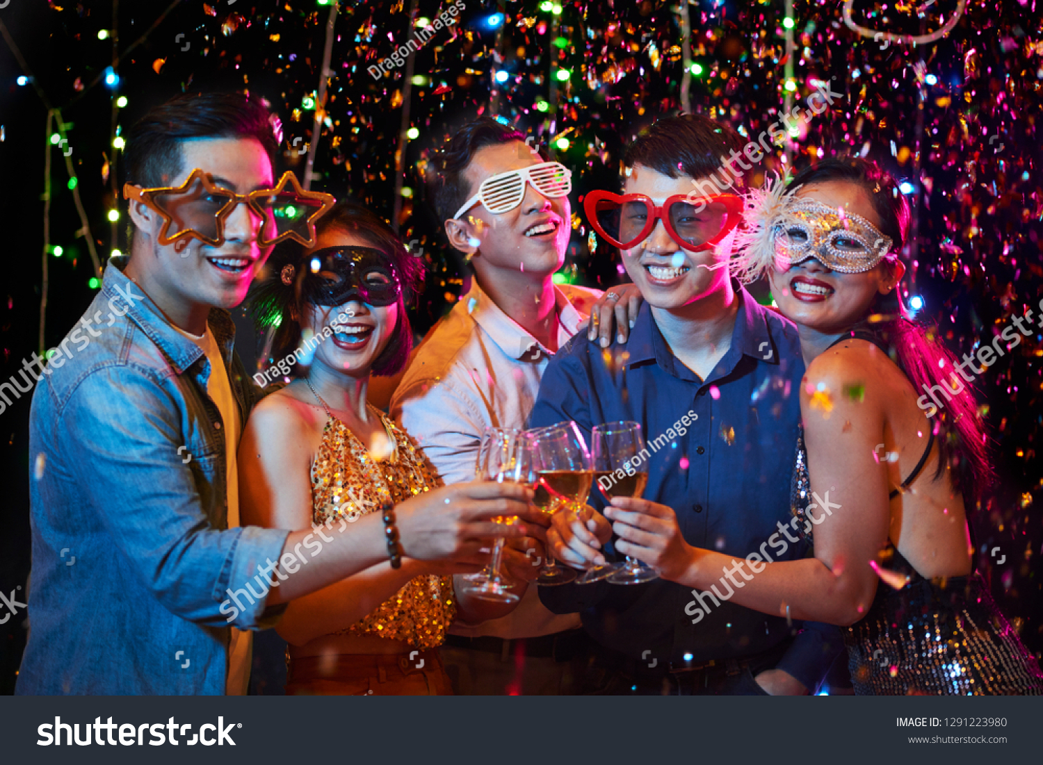 Happy excited young Vietnamese people in party glasses posing with champagne glasses under fallling confetti
