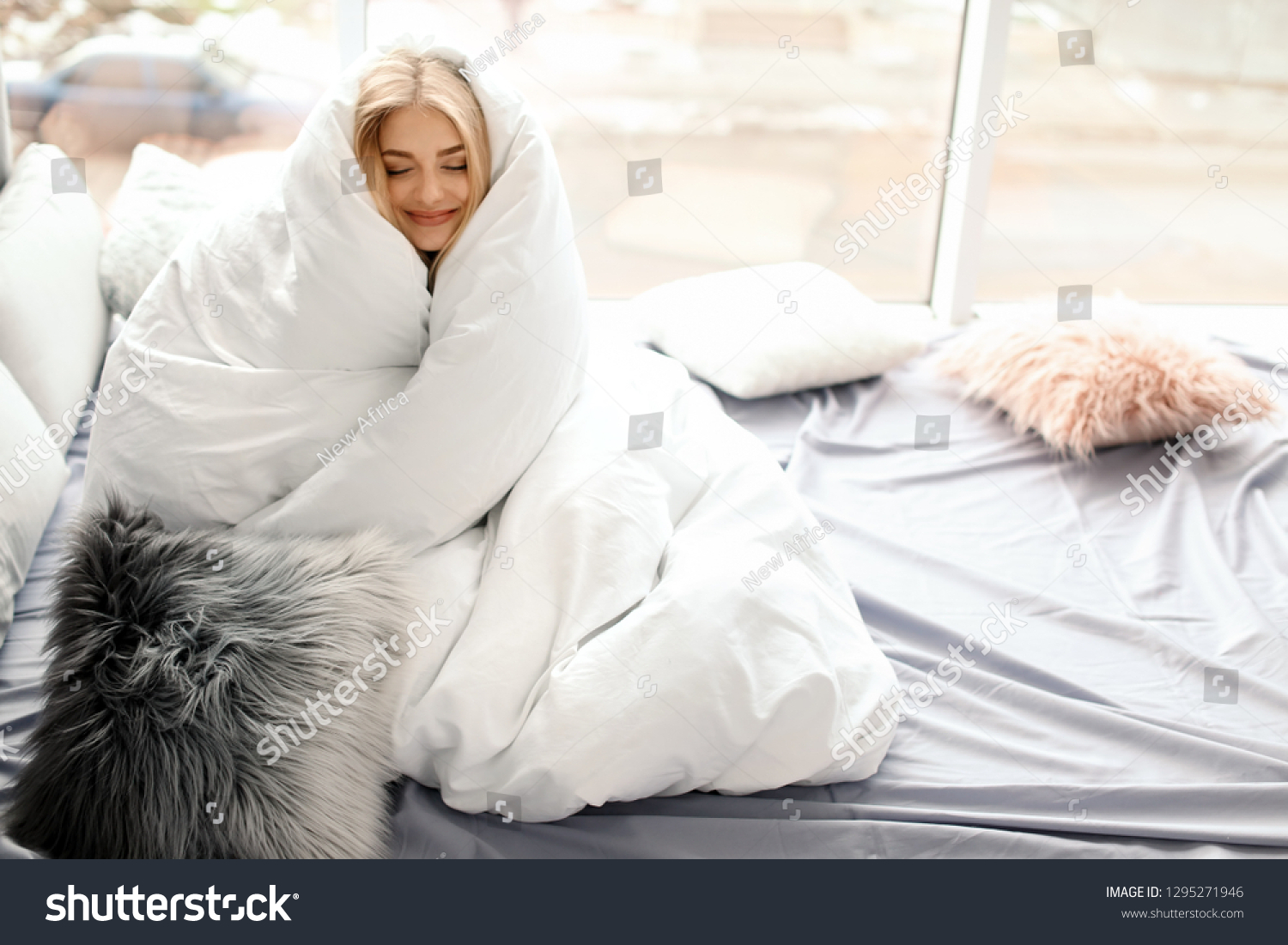 Beautiful young woman wrapped in soft blanket sitting near window at home. Winter atmosphere