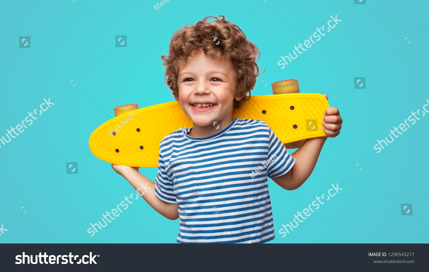 Charming curly boy holding yellow pennyboard and looking away isolated on aqua blue background.
