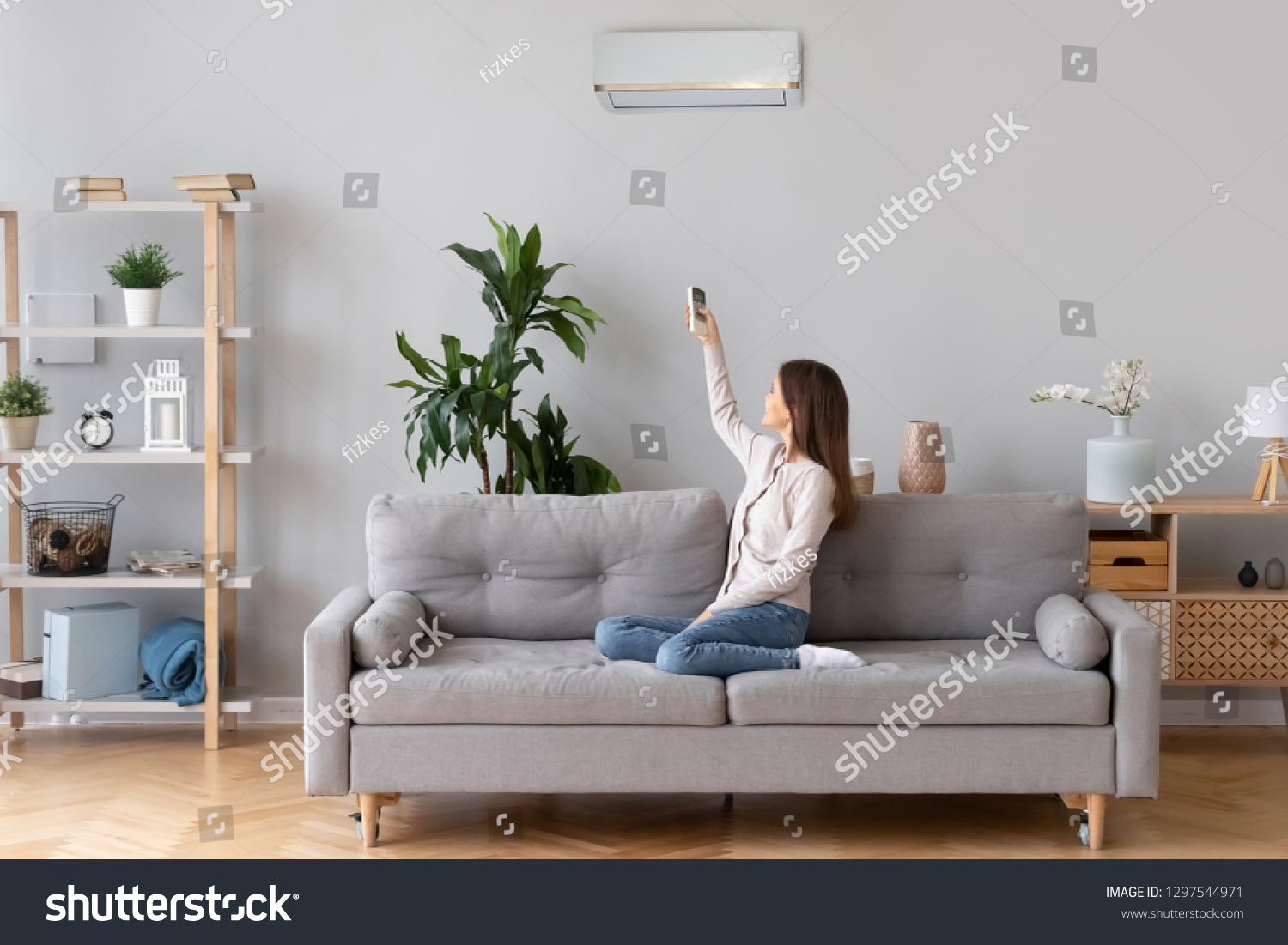 Young happy woman switching on air conditioner sitting on couch at convenient cozy home lady relaxing on sofa in living room holding remote climate control to cooler system set comfort temperature