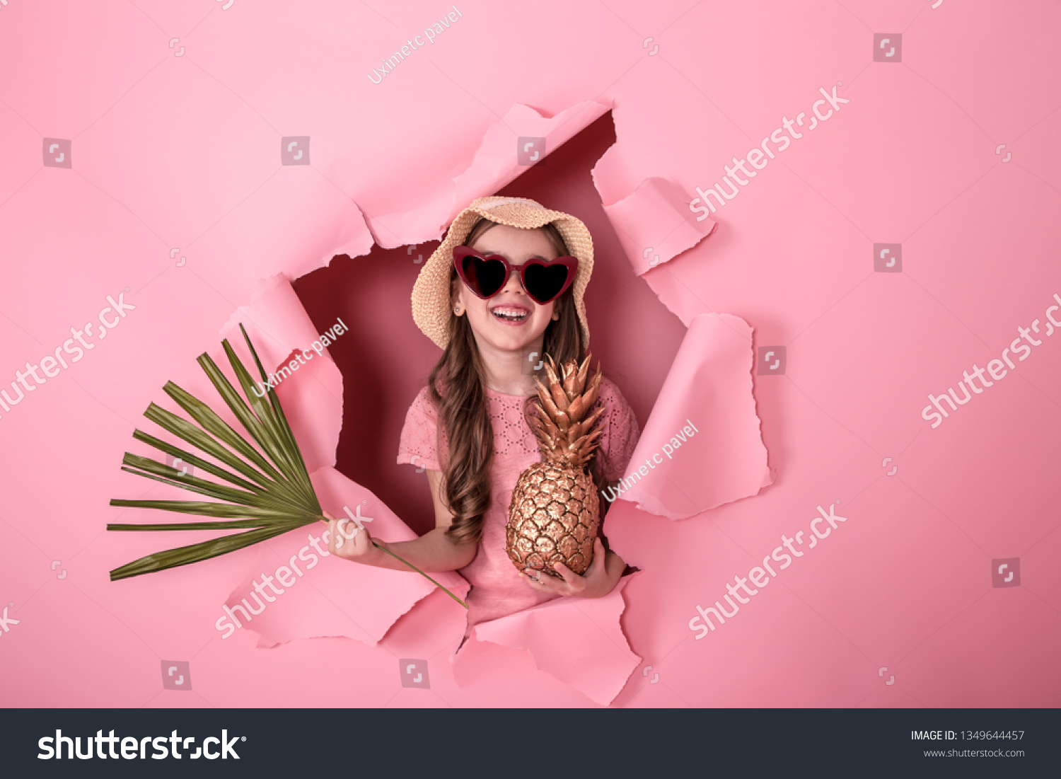 funny little girl peeking out of a hole in a beach hat and heart-shaped glasses with pineapple in her hands and tropical leaves  on a colored background  space for text  Studio shooting
