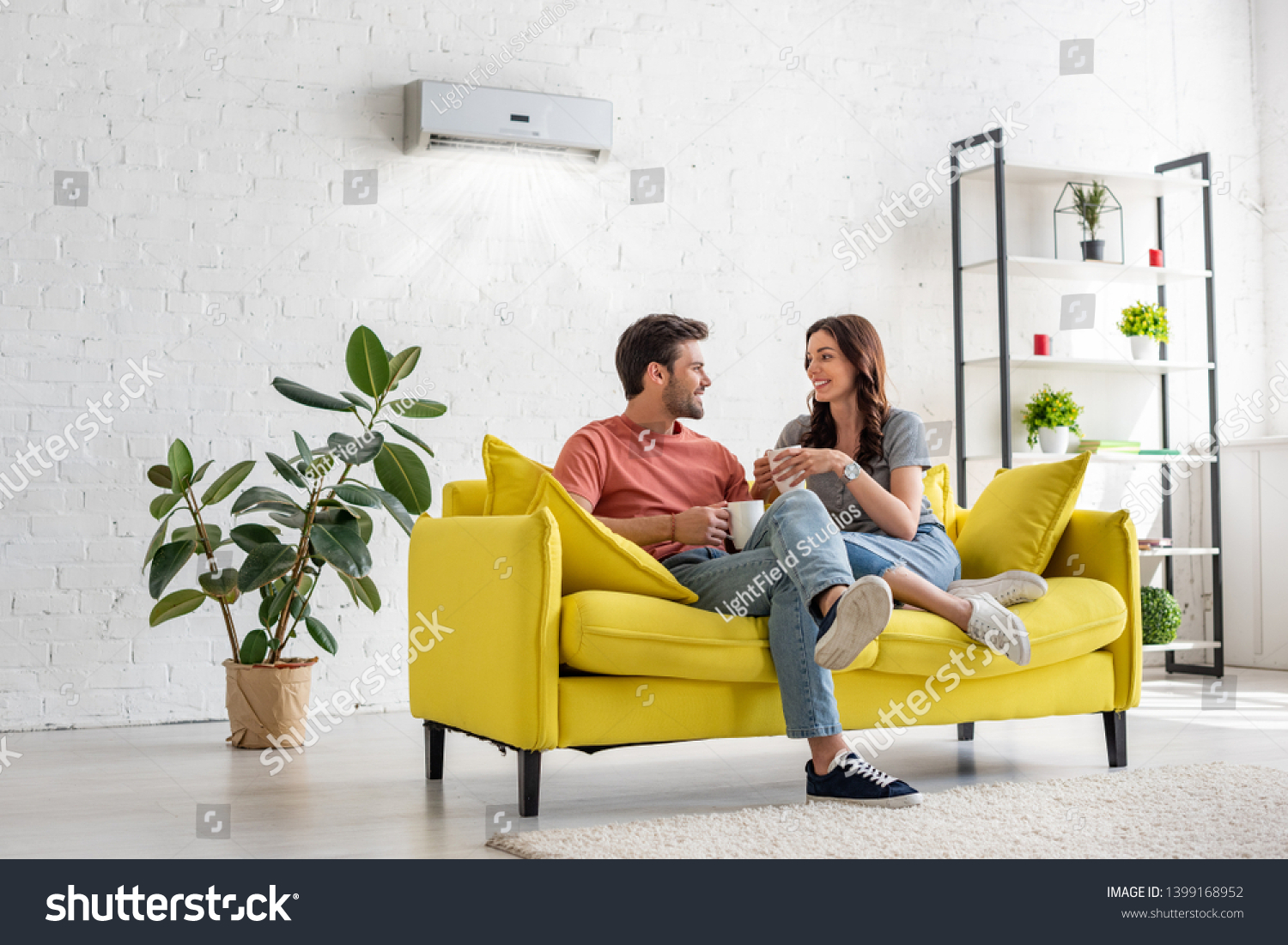 happy young couple talking and holding cups while sitting on yellow sofa under air conditioner at home