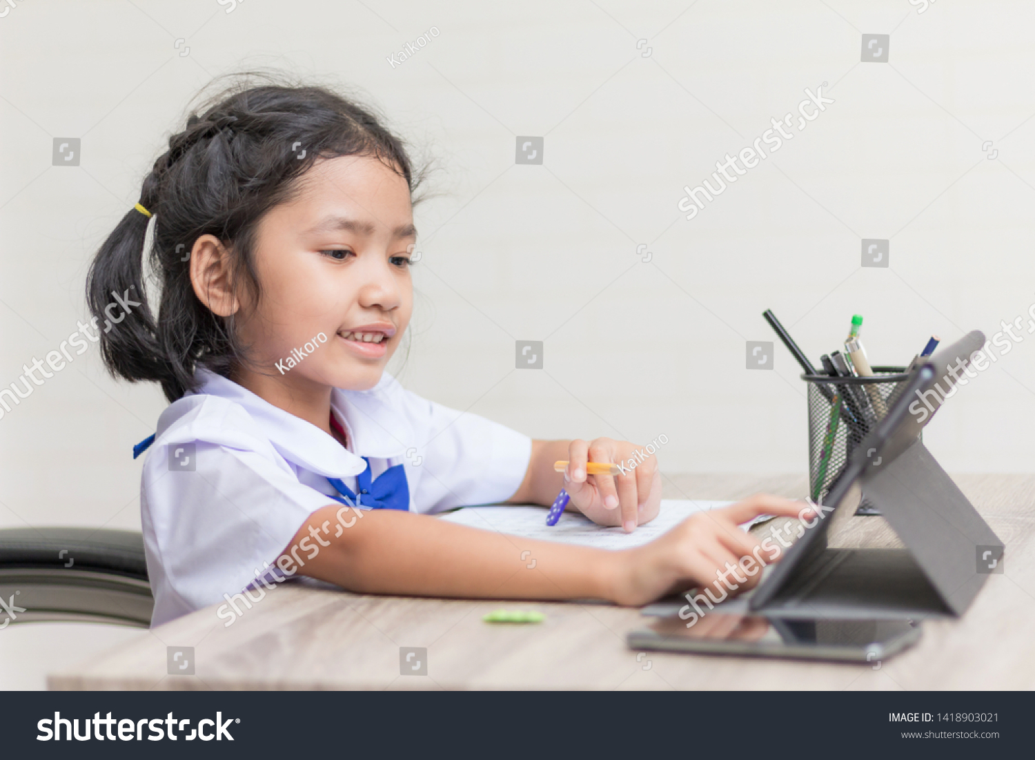Asian little girl in student uniform doing homework and using tablet on wooden table select focus shallow depth of field