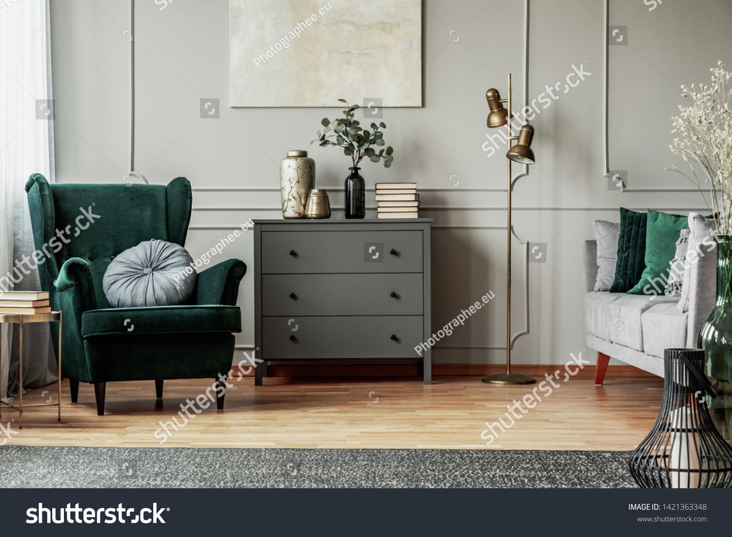 Abstract painting on grey wall o contemporary living room interior with emerald green armchair with round pillow commode and couch