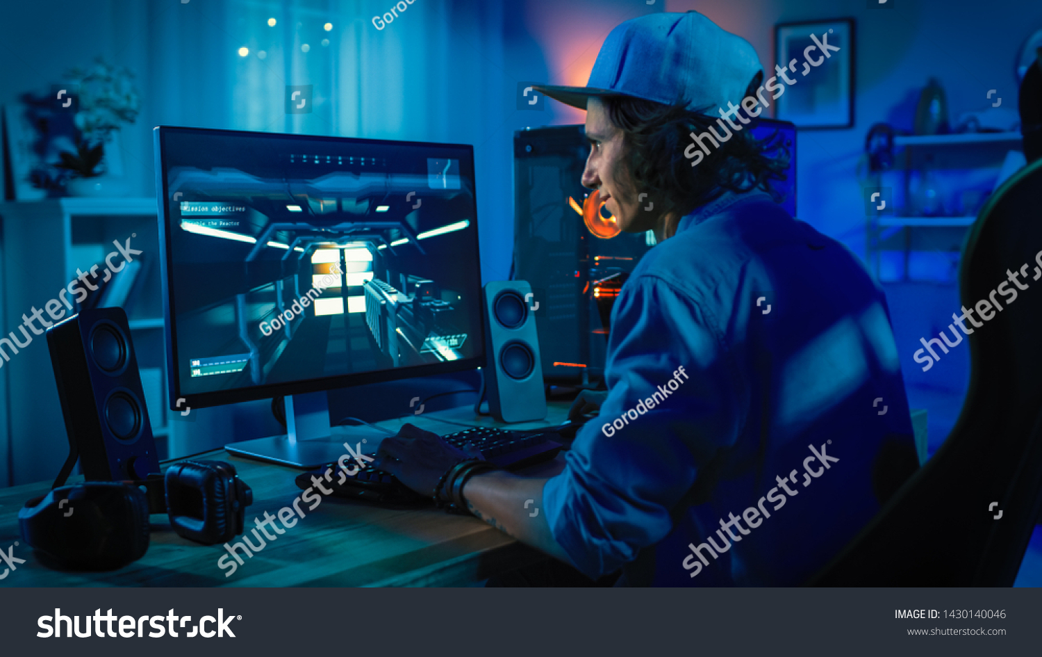 Professional Gamer Playing First-Person Shooter Online Video Game on His Powerful Personal Computer with Colorful Neon Led Lights. Young Man is Wearing a Cap. Living Room Lit in Low Key Style. Evening