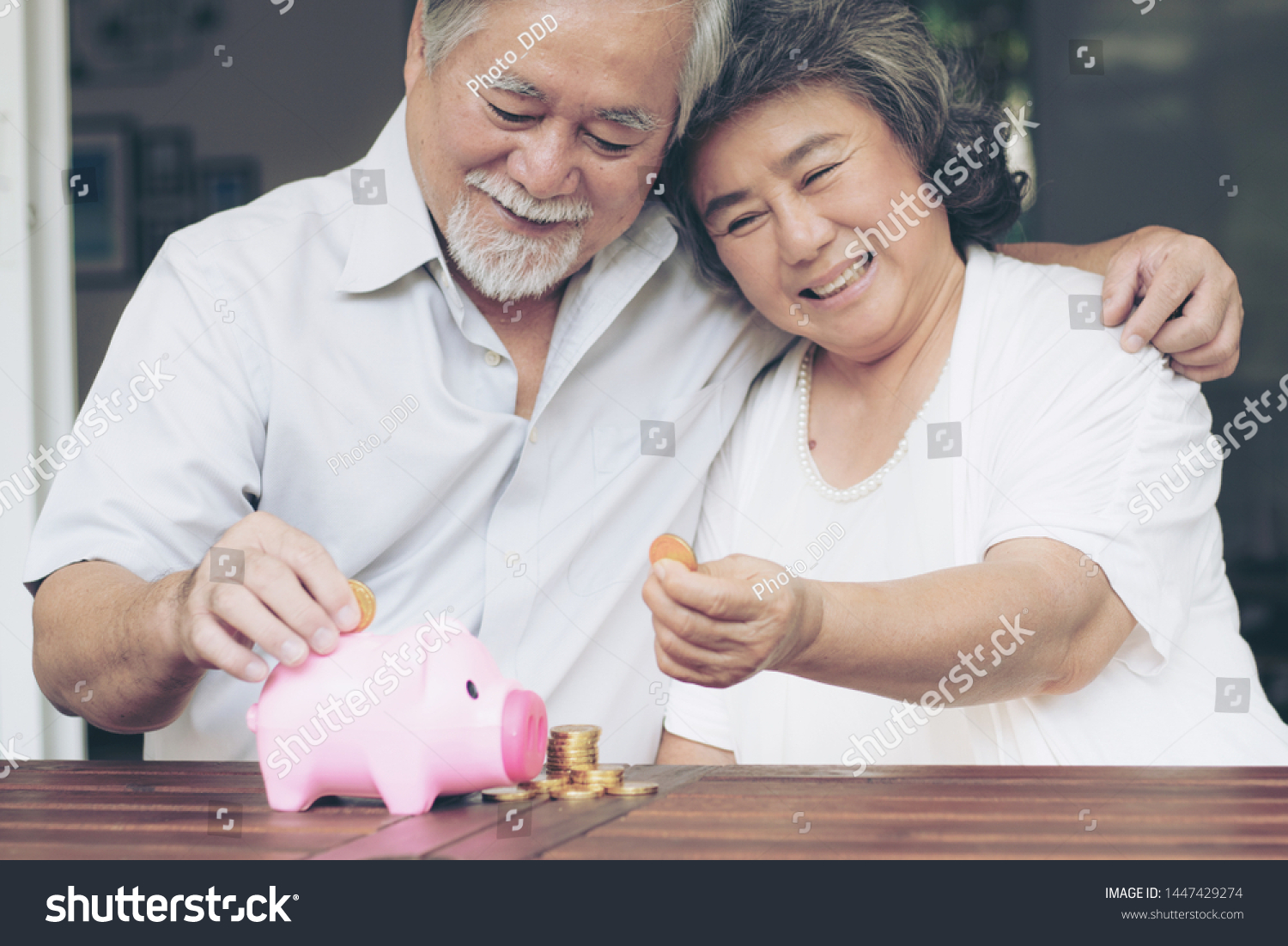 Cheerful elderly couple saving money with putting coin into piggy bank and stack of coin on wooden table They are smiling with happiness together
