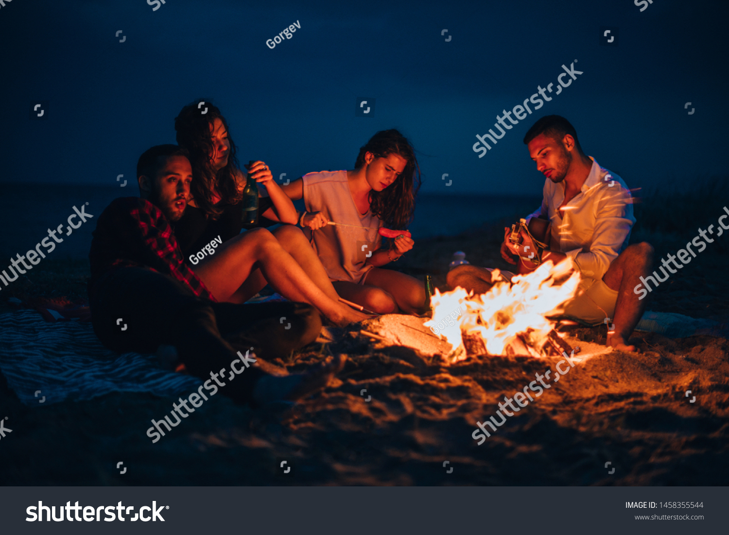 Young couple enjoying with friends around campfire on the beach at night by the sea while listening to a guitar songs