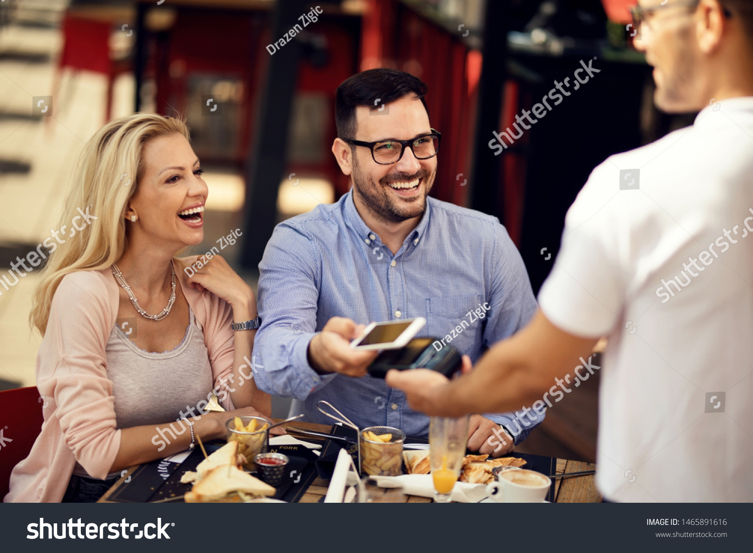 Happy couple having fun while making contactless payment via smart phone in a bar. 
