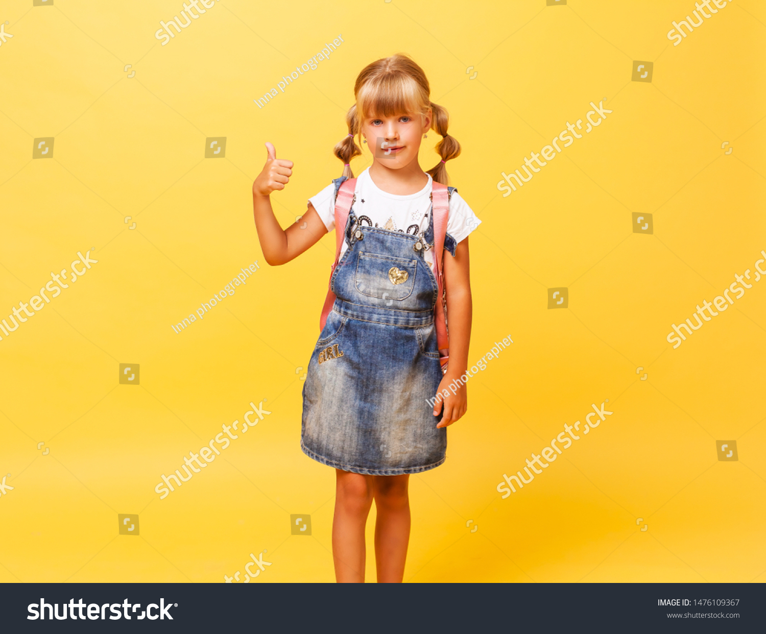 Happy little girl with blond hair and with pink backpack jumps in jeans overalls on a yellow background. Jeans denim.