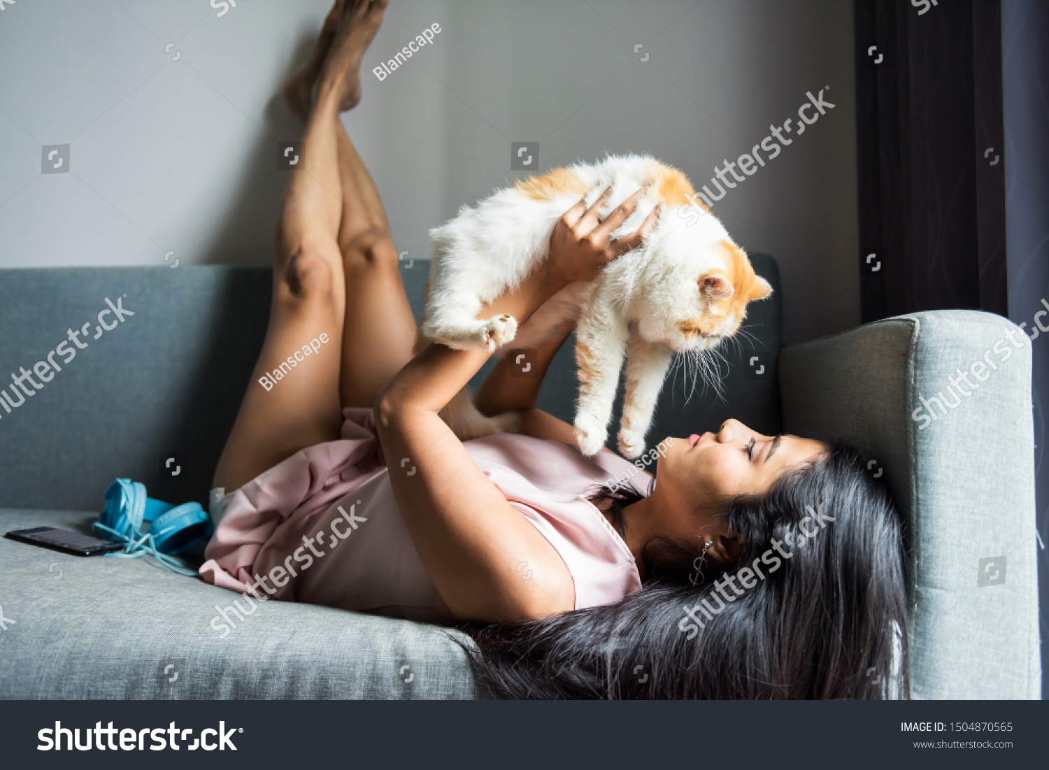 Beautiful Asian girl holding and cuddling cute yellow Exotic shorthair cat on sofa in condo living room. Woman playing with pet in living room at weekend.