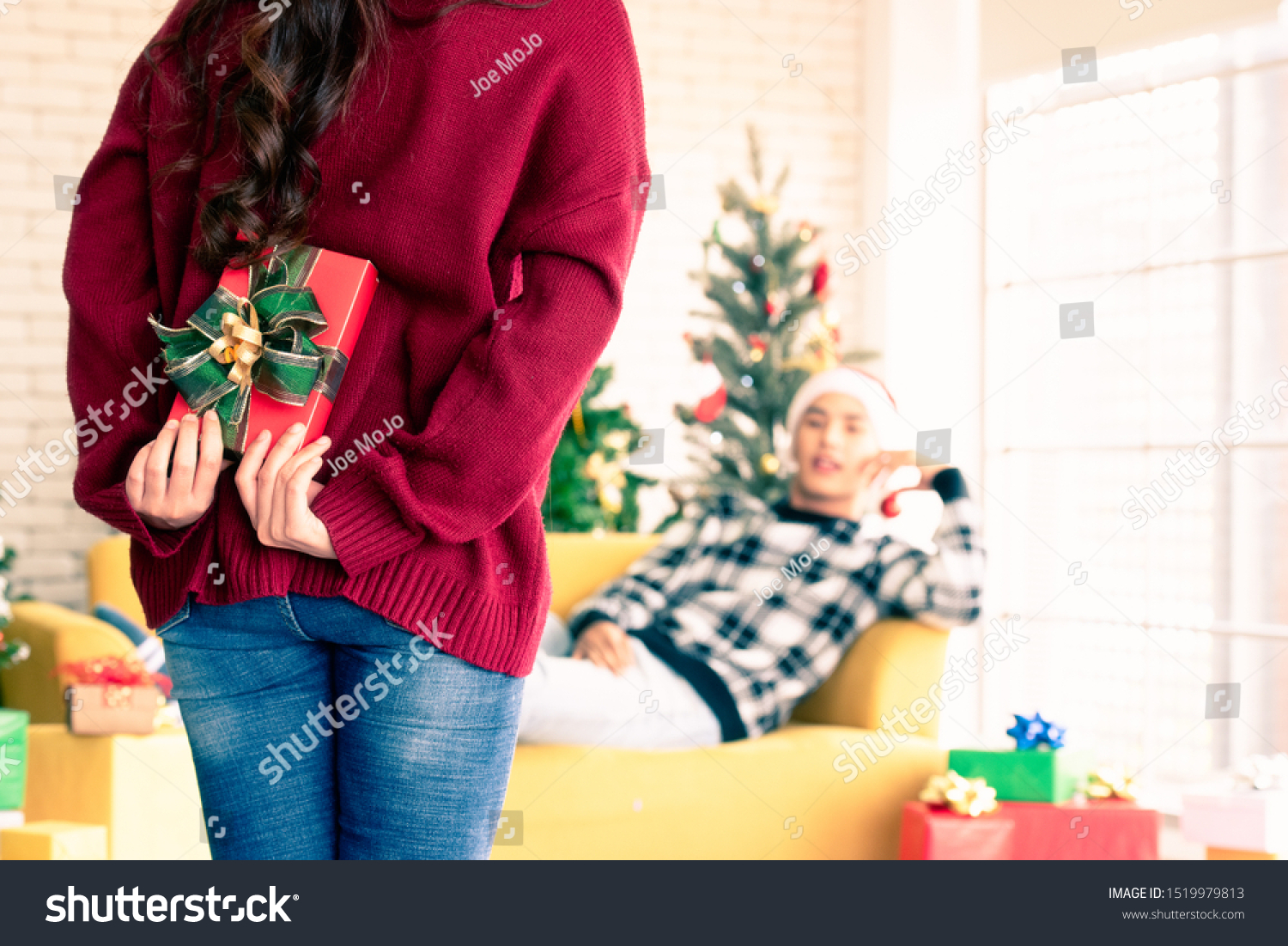 A beautiful Asian woman is surprising his boyfriend by hiding a gift box in the back and walking towards him while the man is lying on the sofa in the room with a Christmas tree behind them.