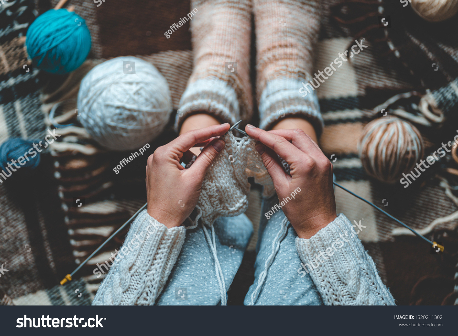 Cozy knitting woman in knitted winter warm socks and in pajamas enjoys knit work on brown checkered plaid blanket at home in cozy winter time. Top view