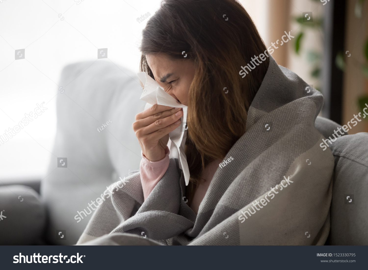 Ill upset young woman sitting on sofa covered with blanket freezing blowing running nose got fever caught cold sneezing in tissue sick girl having influenza symptoms coughing at home flu concept