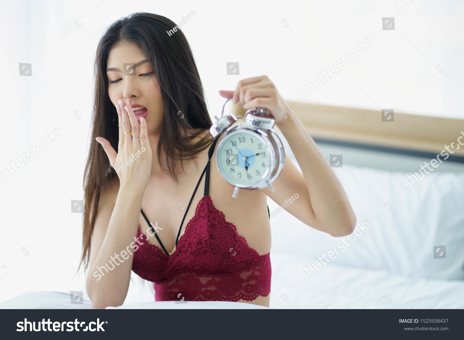 Portrait of sleepy asian woman in sleepwear yawning after her waking up so early in the morning Sitting on bed and holding alarm clock.