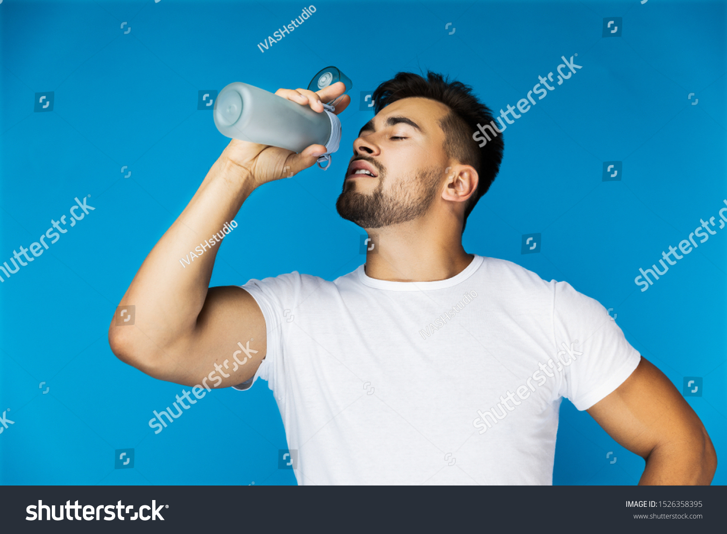 thirsty athletic handsome guy in a white shirt on the blue background is drinking from the sport bottle