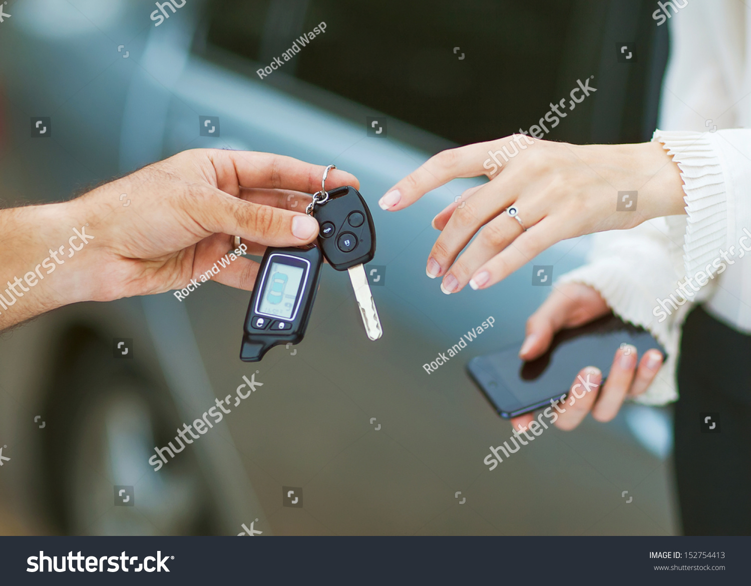Male hand giving car key to female hand. She is holding a cell phone. In the background a fragment of the car.